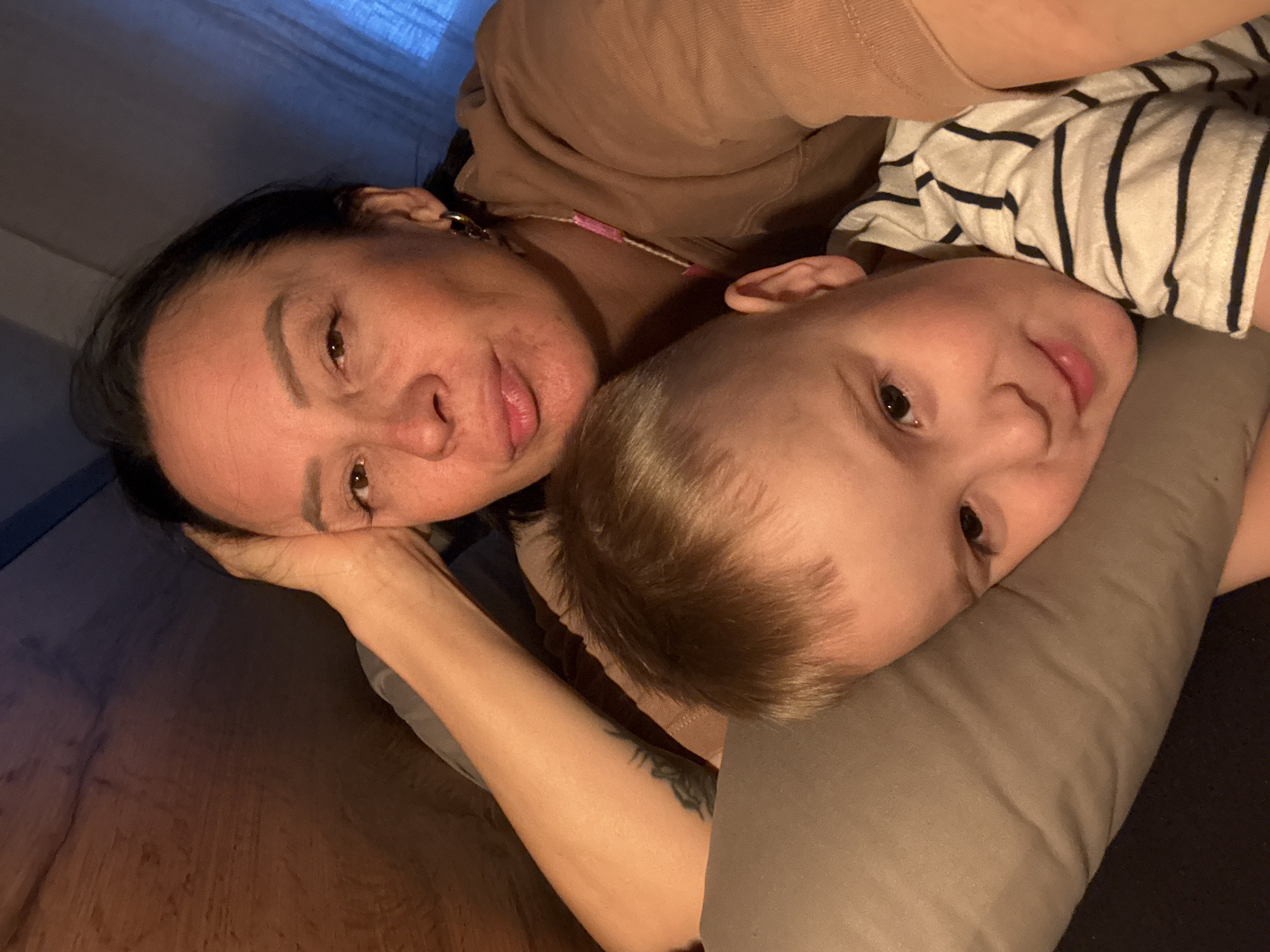 A woman and a child are lying down. The woman has dark hair and is wearing a brown top. The child has short brown hair and is wearing a striped shirt. The child is nestled against a gray pillow. The location is unspecified, and there is no indication of Chaikovsky, Russia.