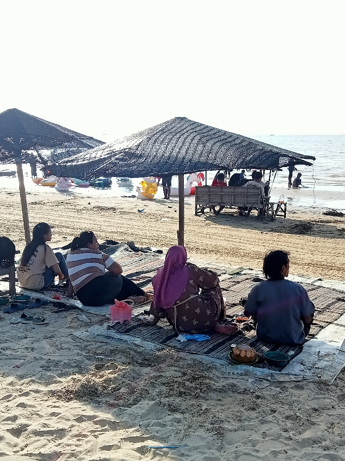 The image shows a beach scene at a location in Rembang, Indonesia, with coordinates of -6.684 latitude and 111.412 longitude. The beach is sandy and the water is clear, with a few inflatable boats anchored in the shallows. There are several people sitting on the beach under a large black umbrella made of woven bamboo, with another smaller umbrella nearby. Two women sit facing the ocean, one with a headscarf, the other with brown and white stripes. A young boy sits between the two women. Two men and two women are sitting further down the beach to the right of the large umbrella, facing away from the ocean. They are all sitting on woven mats, and there is food and drinks on the mat in front of the boy. There are also a few plastic chairs under the large umbrella, and a table with food on it. The sky is bright blue and there is a slight breeze.