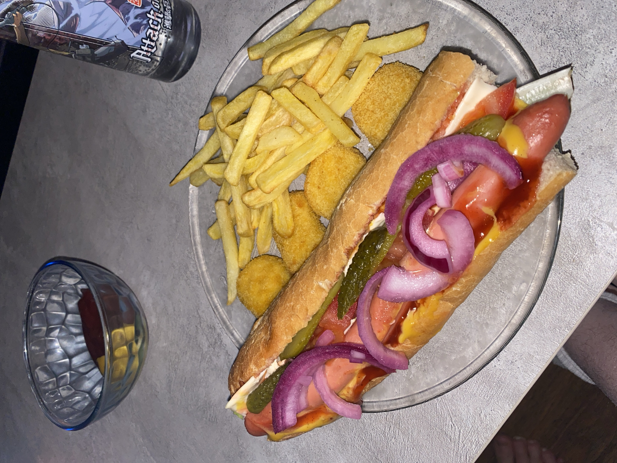 A high-angle shot depicts a clear glass plate resting on a light grey surface, containing a loaded hot dog, french fries, and fried spherical appetizers. The hot dog is split in a baguette-style bun and topped with what appears to be ketchup, mustard, mayonnaise, tomato slices, pickle slices, and red onion rings. Adjacent to the hot dog are a serving of golden-yellow french fries and a cluster of golden-brown, round fried appetizers. To the left of the plate sits a glass bowl containing a reddish sauce or liquid. A dark bottle with branding in a language using non-Latin characters is partially visible in the upper left corner of the image. A portion of a person's leg and foot are visible in the lower right corner. The surface appears to be a countertop or table, with a glimpse of a wooden floor underneath. There is no information indicating the image was taken in Izhevsk, Russia.