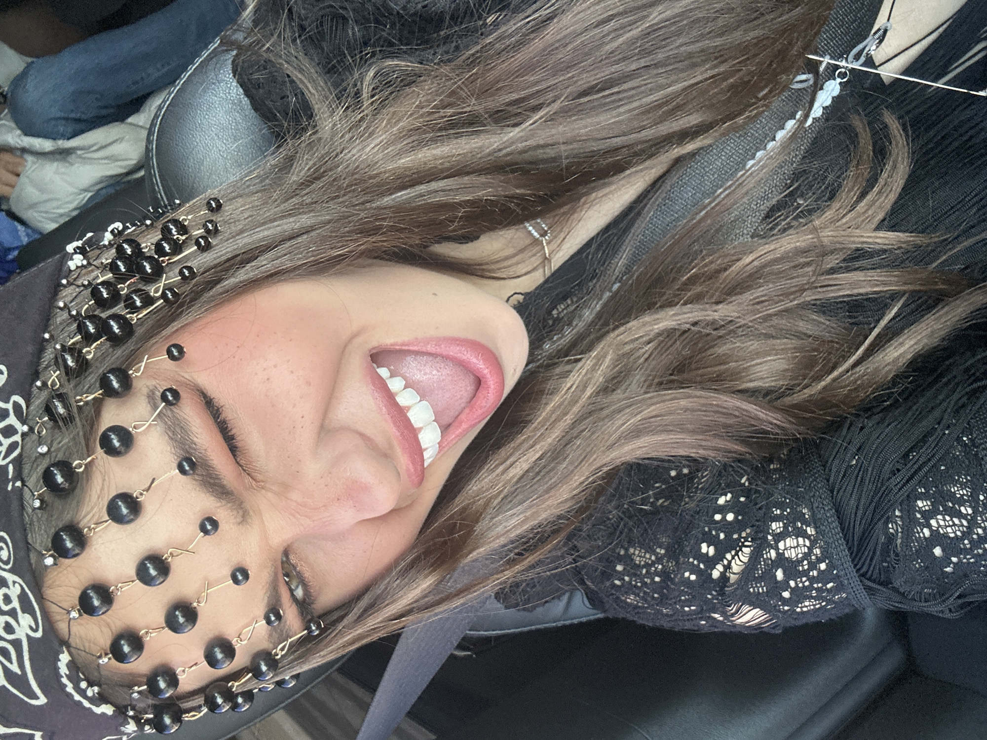 A woman is seen at an unusual angle, possibly lying down in a vehicle. She has fair skin, light brown hair, and is wearing a decorative headpiece of black beads. She has her mouth open, displaying her teeth and tongue. She appears to be wearing a black top with lace or fringe details. A gray seatbelt is visible across her body. The environment is indistinct, but the scene suggests an indoor setting, potentially inside a car. The location is assumed to be Palm Desert, United States of America.