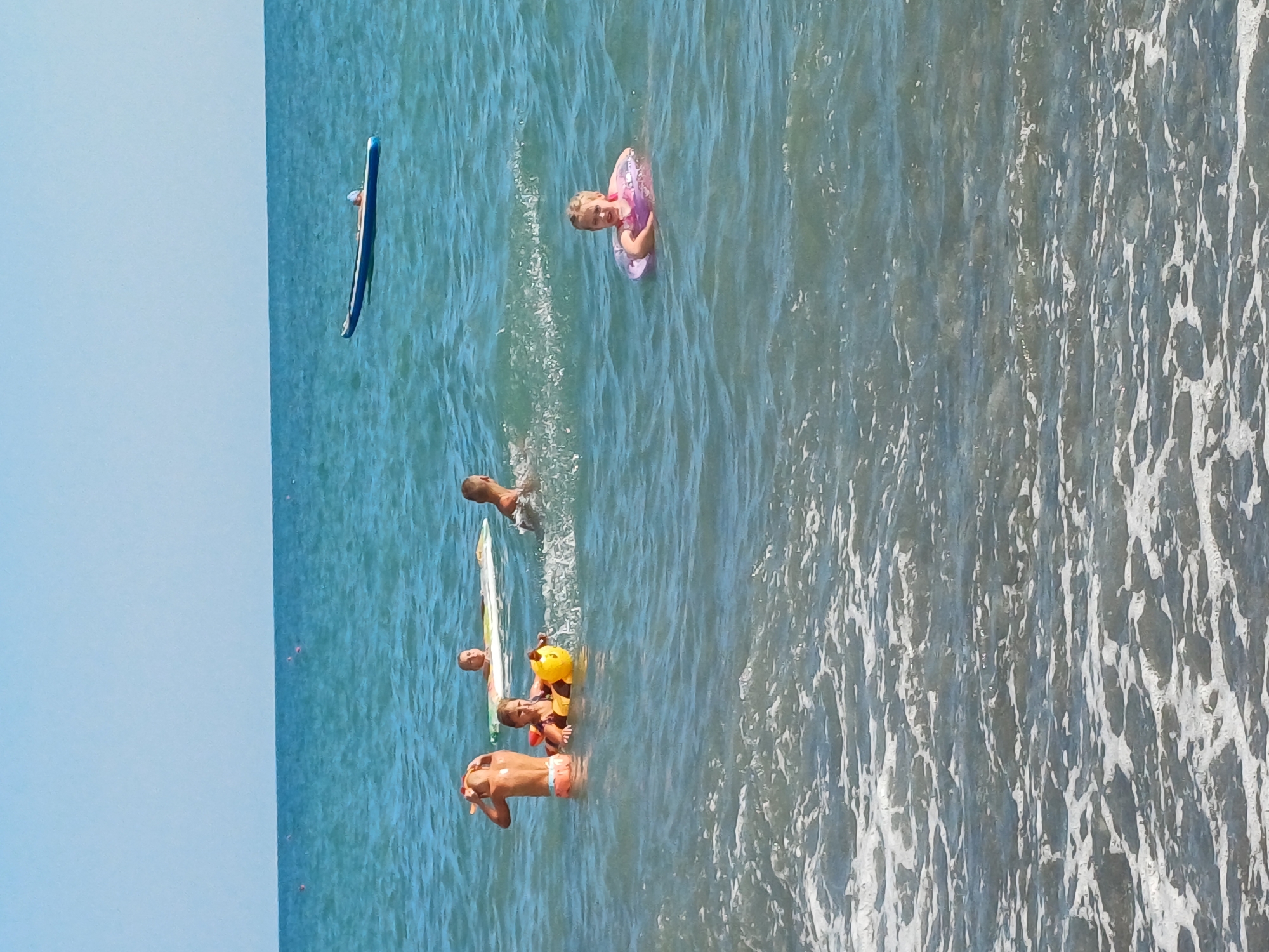 The image depicts a sunny day at the Black Sea coast in Sochi, Russia, with several children enjoying the water. In the foreground, a young girl with blonde hair is swimming with a pink inflatable ring around her. To the left, a boy wearing orange swim trunks is partially submerged, looking towards the left side of the frame. Next to him, another child is seated on a yellow inflatable toy shaped like a bee. Further in the background, two other children are visible: one on a white inflatable mat, and another swimming. In the mid-ground, a long, blue and white inflatable paddleboard is floating on the water. The sea is calm with small waves and a clear blue sky overhead. The shoreline is not visible in this image, as the focus is on the water activities.