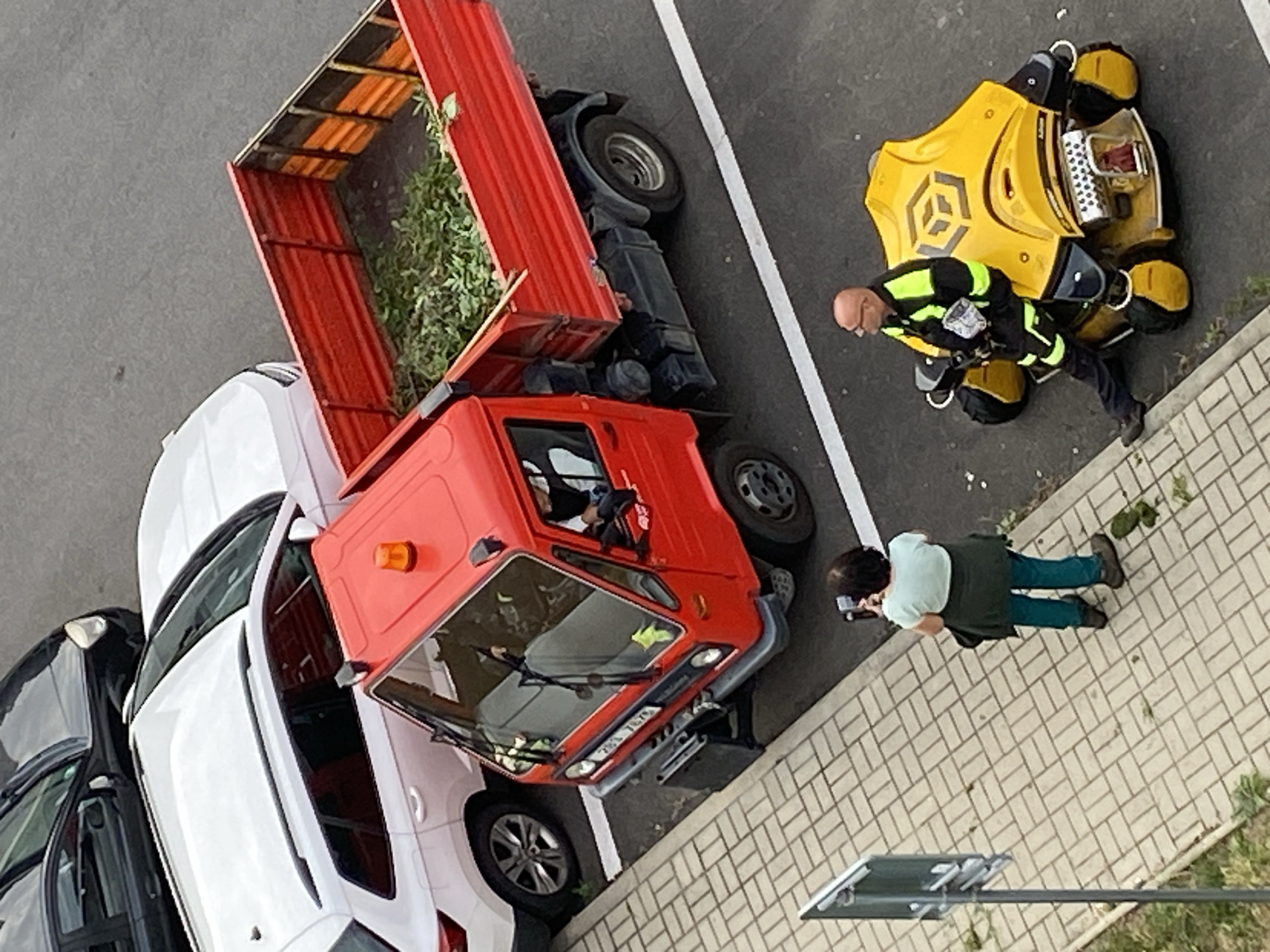 In Bílina, Czechia, on an asphalt surface next to a brick-paved sidewalk, a red dump truck filled with green branches is parked. Adjacent to the truck, a person is taking a photograph with a camera. Further down the sidewalk, a man in a high-visibility jacket is standing next to a yellow ride-on machine. Two cars, a white SUV and a black car, are parked behind the dump truck.