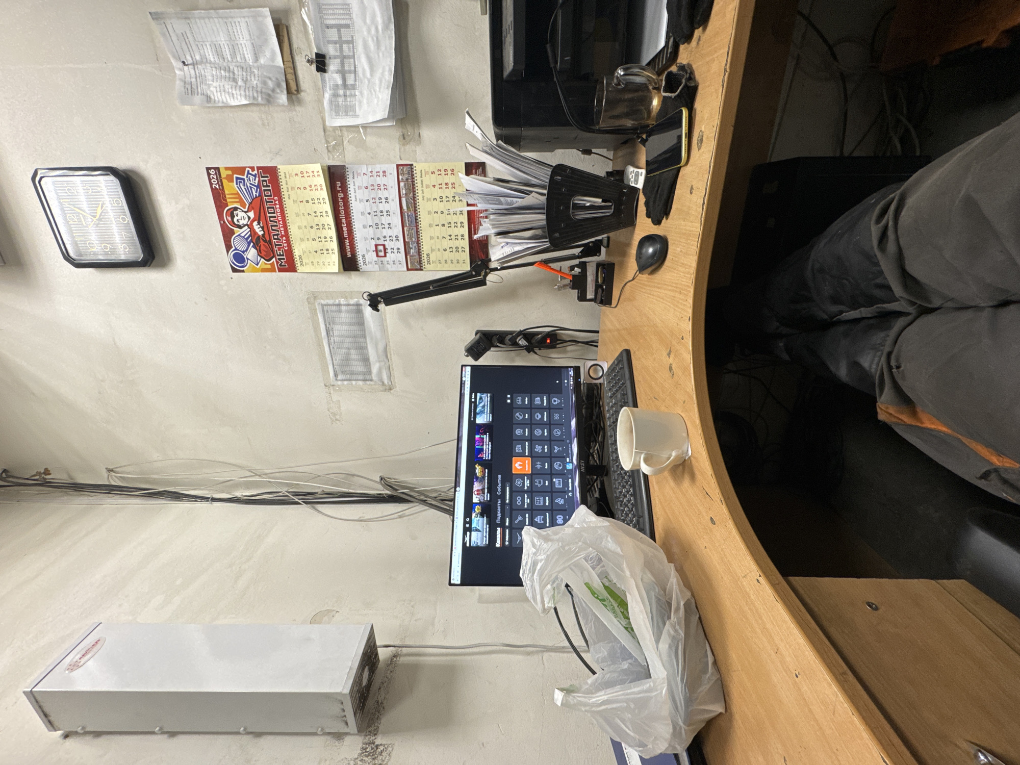 The image depicts an office workspace with a desk, computer, and various office supplies. The desk is made of wood and has a curved shape. On the desk, there is a computer monitor displaying a menu with icons, a keyboard, a mouse, a white mug with a tea bag string hanging out, a plastic bag containing some items, a pen holder filled with papers, a printer, a glass mug with liquid, and a smartphone. In the background, there is a wall with several calendars and papers attached. A white rectangular device is mounted on the wall to the left. The scene appears to be an indoor office environment. The image does not provide any specific details about the city of Sakmary, Russia, or its location within it.