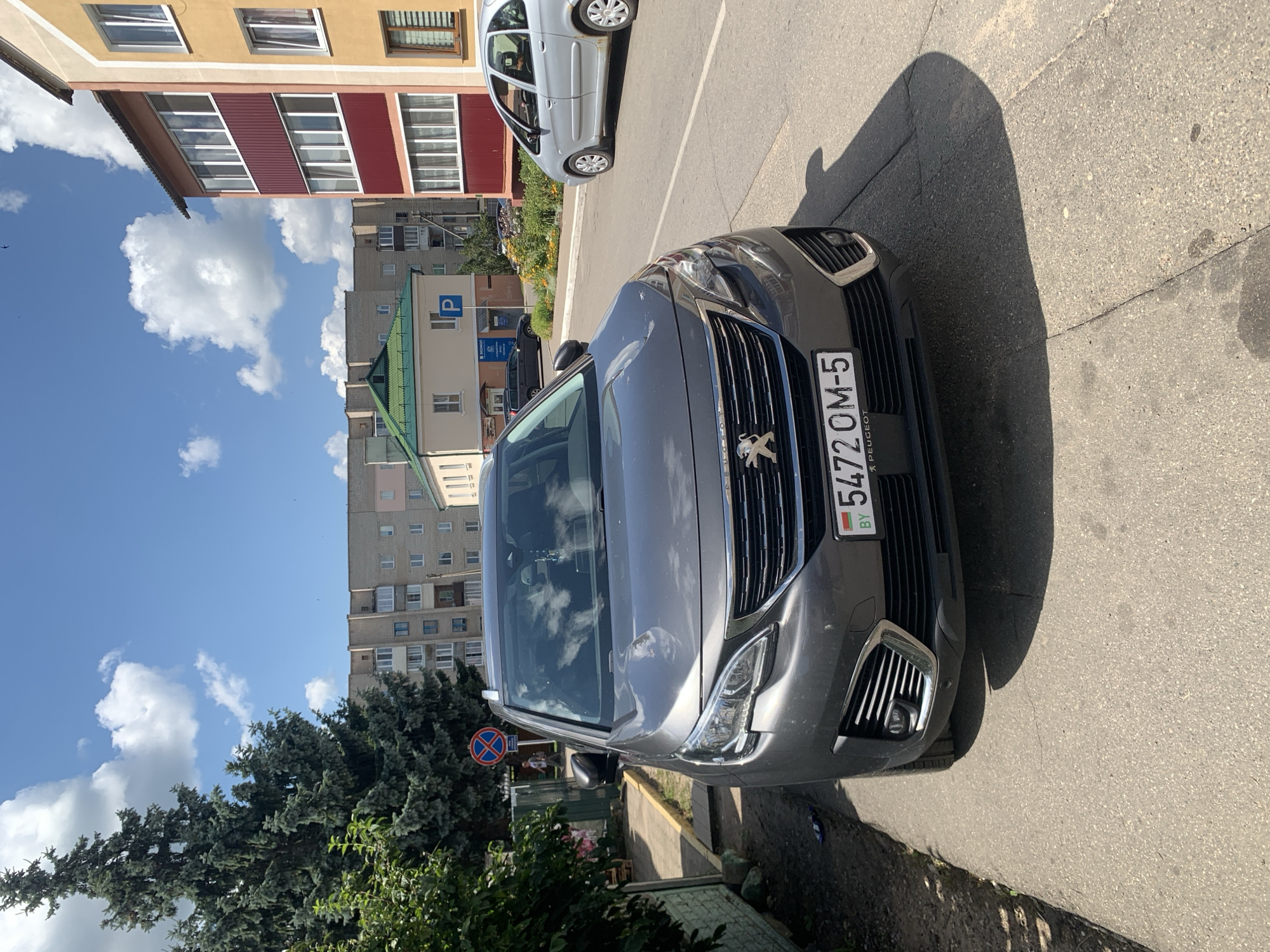 A gray Peugeot 5008 SUV is parked on an asphalt road in the city of Kapyĺ, Belarus. The car is positioned facing the viewer, with its front bumper and grille prominently visible. The license plate reads "5472 OM-5" with "BY" on the left, indicating Belarusian registration. A tall evergreen tree stands to the left of the car, casting a shadow. Behind the SUV, several apartment buildings with balconies are visible. A light-colored sedan is parked further down the road. The sky is blue with scattered white clouds. A "no parking" sign is posted near the tree. The scene appears to be a residential area on a sunny day.
