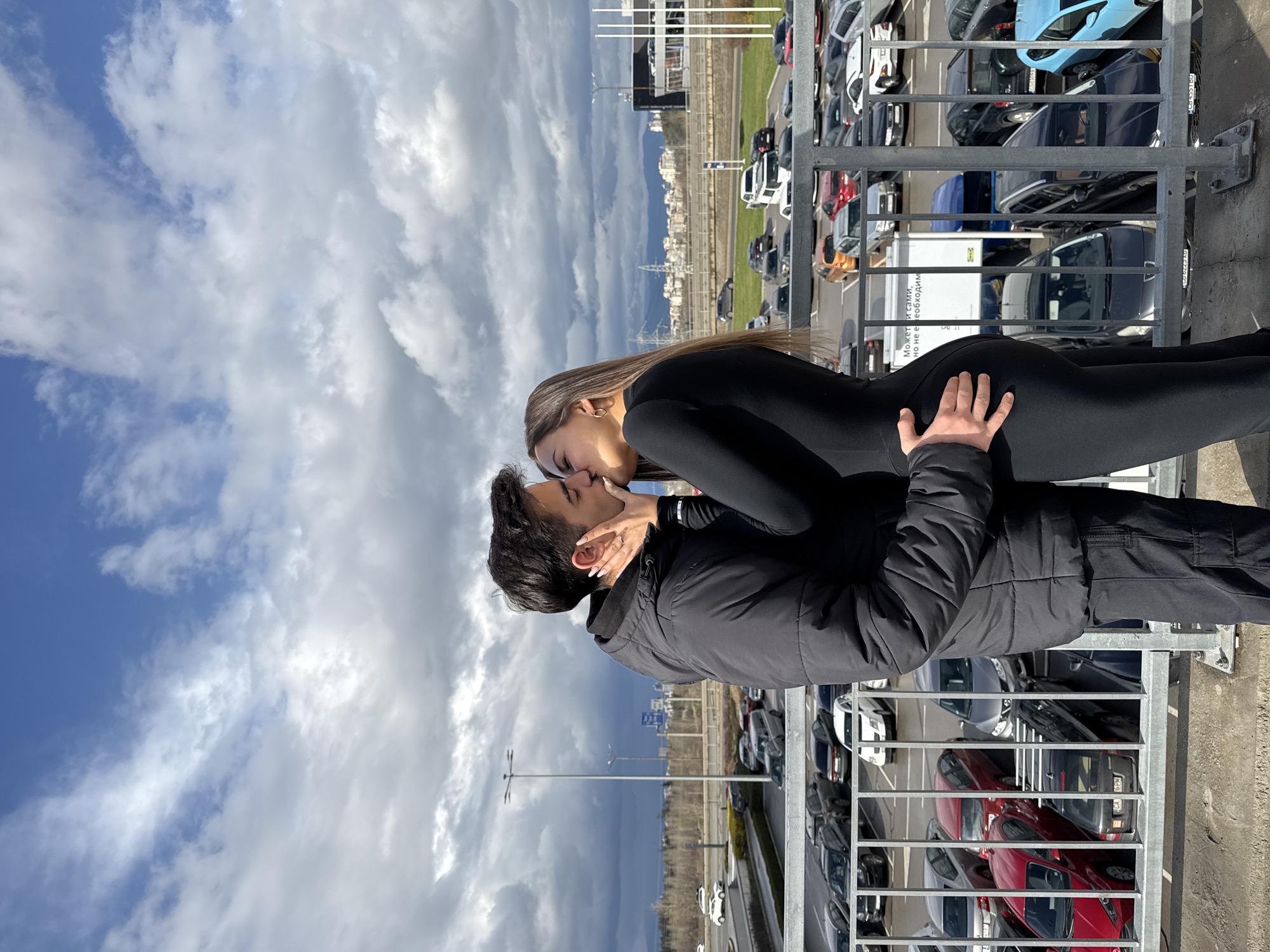 A couple is kissing on a raised platform with a metal railing, overlooking a parking lot filled with various cars. The woman is wearing a black long-sleeved bodysuit and the man is wearing a black jacket. The man's hand is on the woman's backside. In the background, there is a cityscape under a partly cloudy sky. The text on a white sign in the background is in Cyrillic. The location is in Sofia, Bulgaria based on the style of Cyrillic writing.