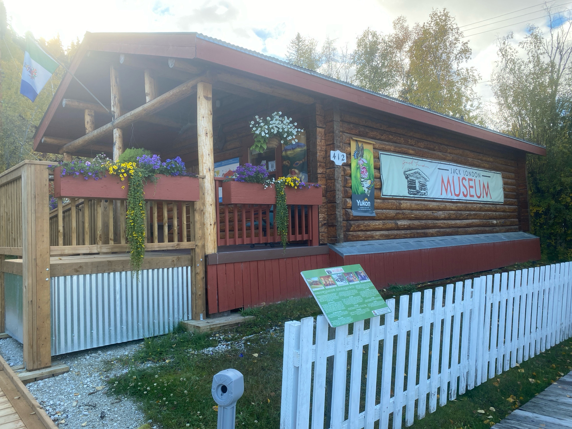 The image shows the Jack London Museum in Dawson City, Yukon, Canada. The museum is a log cabin with a red roof and a wooden porch. There are flower boxes on the porch with purple and yellow flowers. A white picket fence runs along the front of the property. A sign on the building reads "Jack London Museum". Another sign in front of the fence has information about the museum. The sky is cloudy and the trees in the background have autumn foliage. There are no people visible in the image.