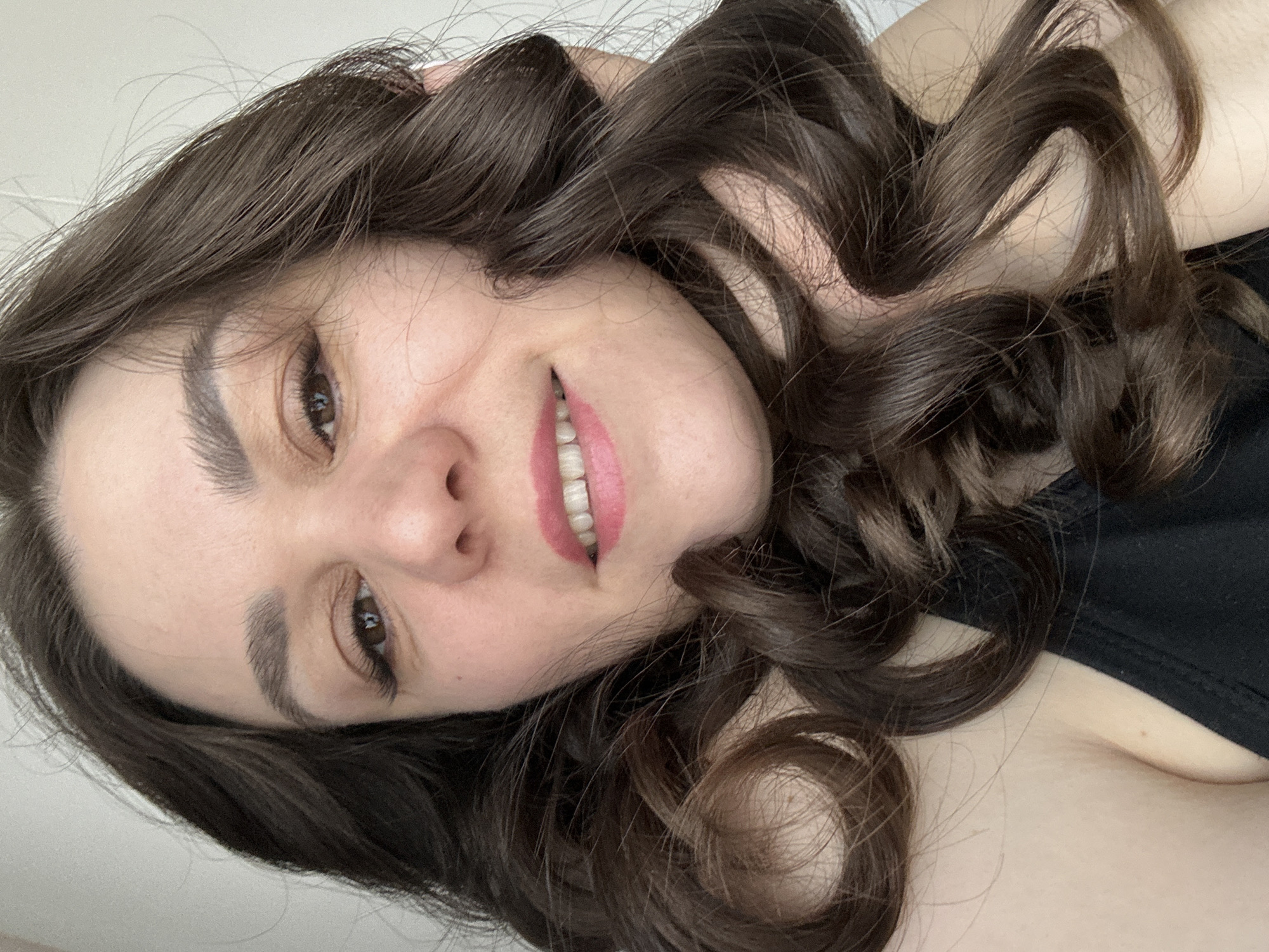 A woman is pictured from a high angle in an indoor setting. She has brown hair styled in large curls and is wearing a dark-colored top. Her face is partially tilted, revealing her eyes, nose, and a smiling mouth with pink lipstick. The background is primarily a plain white wall. There is no indication of the city of Chaikovsky, Russia.