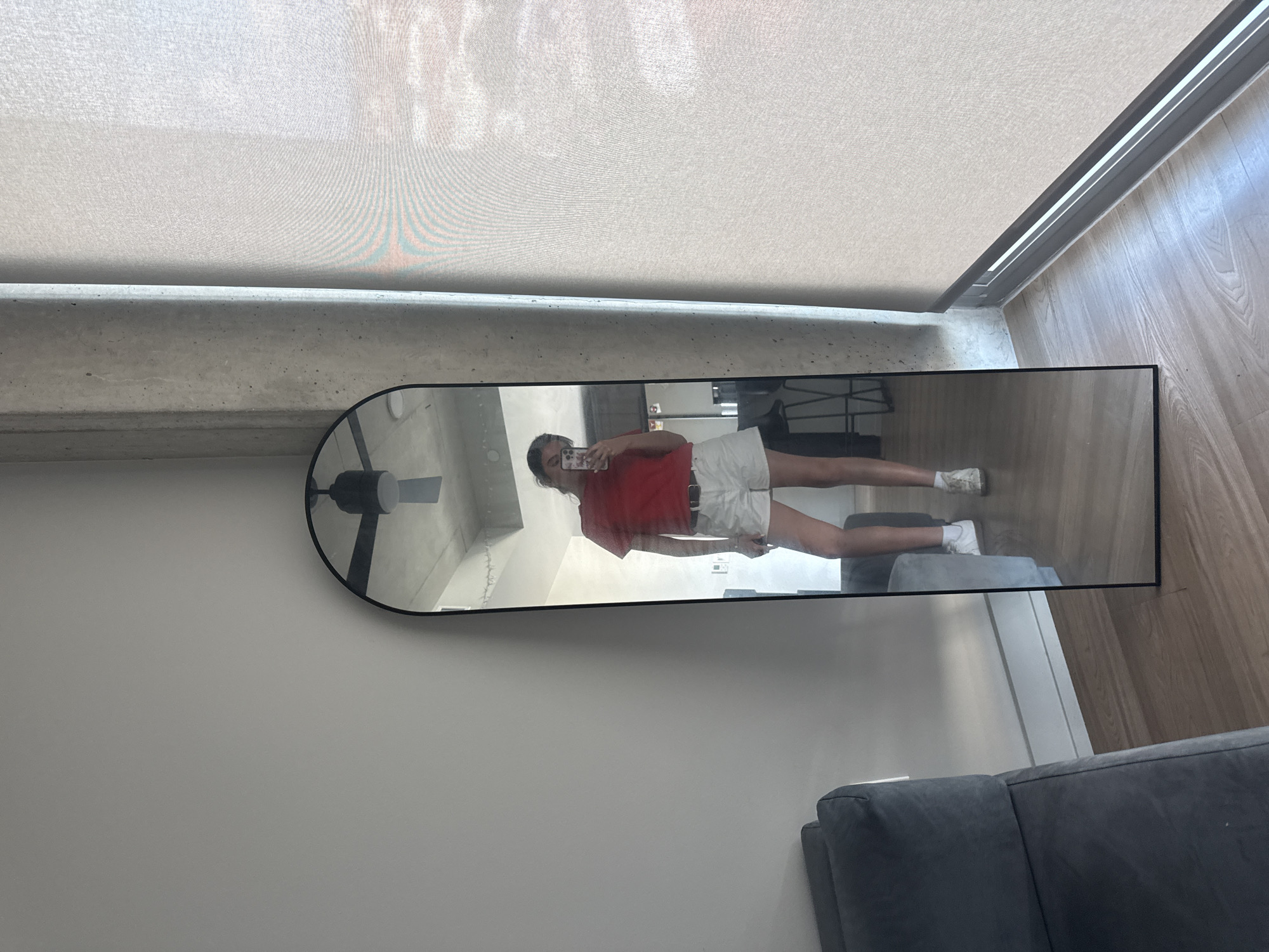 The image is taken indoors, showing a full-length mirror reflecting a person holding a phone, presumably taking a picture of themselves. The person is wearing a red top, white shorts, and white shoes. The reflection also shows a ceiling fan and a partial view of the apartment interior with a sofa. The floor is wooden. The mirror is standing against a white wall, and a window with a shade is visible at the top of the image. The location is inferred to be in Austin, United States of America. No specific landmarks that confirm the Austin location are visible.