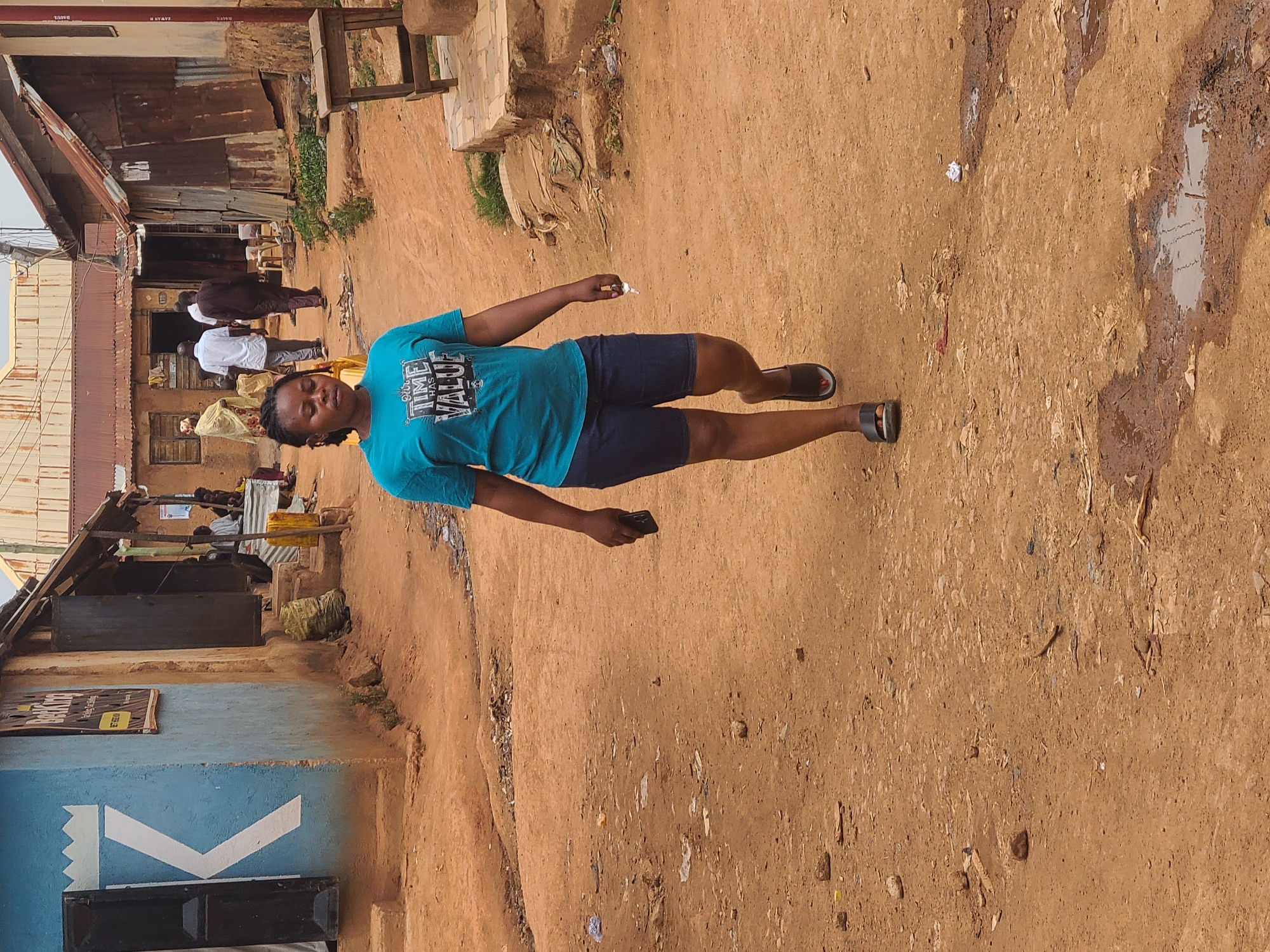 A medium shot shows a woman in a blue t-shirt and navy shorts standing on a dirt road in Jos, Nigeria. She is holding a small white object in her left hand and a black phone in her right. She is looking to the left of the frame. The road is uneven and shows signs of mud and water. Behind the woman are corrugated metal buildings with wooden supports. Several people are walking along the path in front of the buildings. To the left of the woman is a blue building with a white graphic. The lighting is bright and the scene appears to be during the daytime.