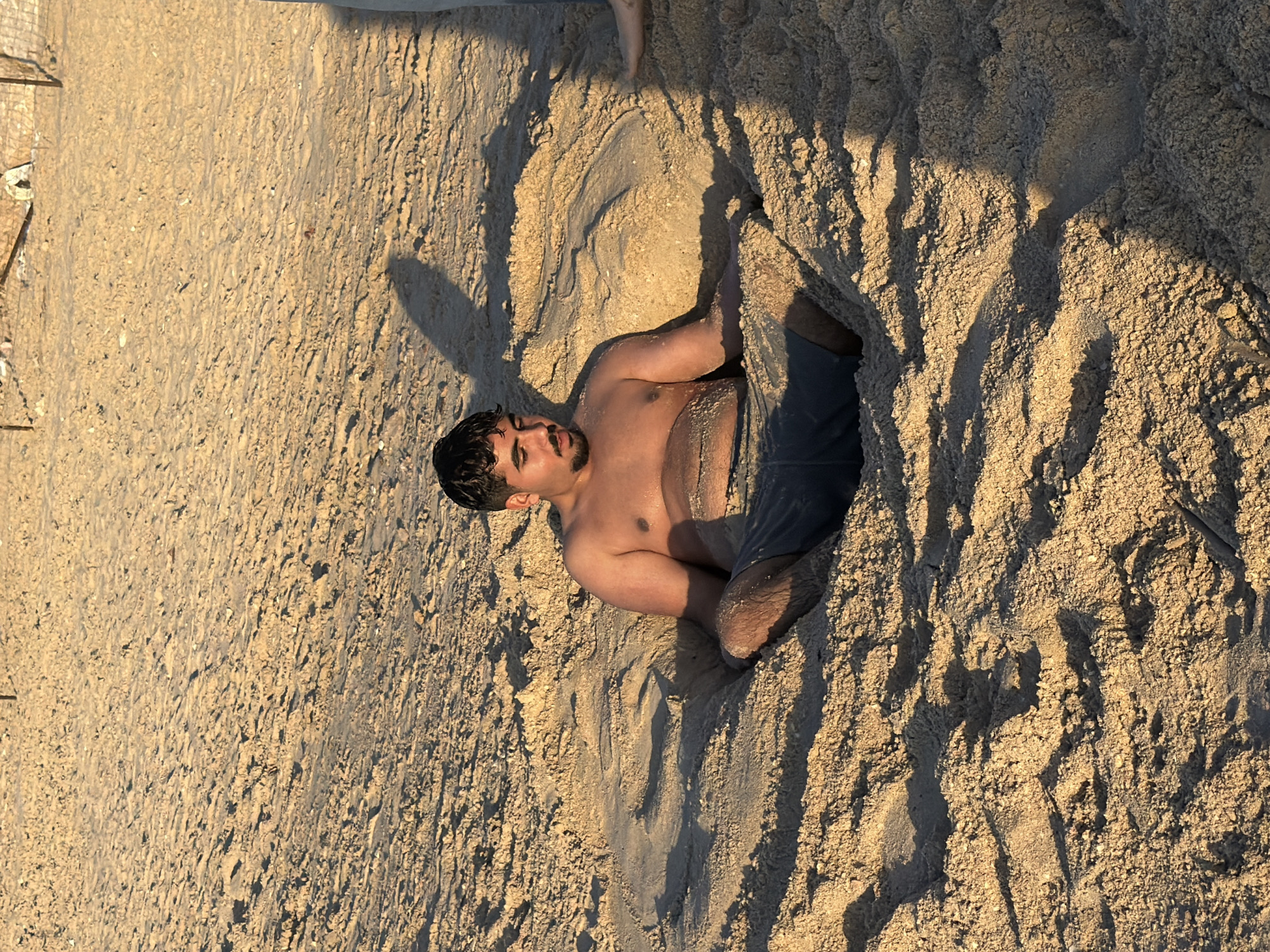 This image captures a man relaxing on a sandy beach, partially buried up to his waist. He is lying on his back, shirtless, wearing dark shorts, with his head turned slightly to the left. He has dark hair and a beard, and his arms are bent, resting on the sand. The man appears calm and content, possibly enjoying a leisurely moment.
The setting is a sunny beach, indicated by the abundance of sand. The lighting is warm and golden, characteristic of the "golden hour" (either sunrise or sunset), casting long, distinct shadows across the textured sand. These shadows are particularly noticeable on the right side of the frame and around the man, emphasizing the contours of the sand mounds. In the upper left portion of the image, a light-colored foot or lower leg of another person is partially visible, suggesting the man is not alone and is likely engaged in a playful activity or simply relaxing with others. The overall scene conveys a peaceful beach environment. No text is visible.