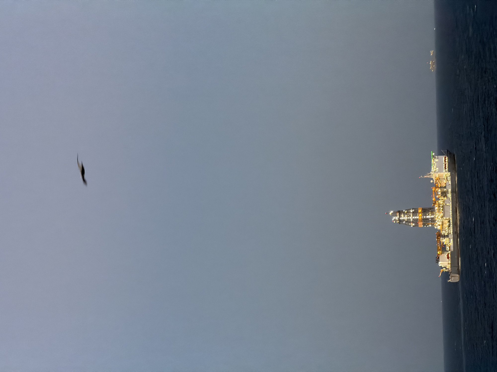 An outdoor scene shows a sideways view of a body of water, presumably the sea, with an oil tanker or similar industrial vessel on the right side, illuminated by many lights. Further in the distance, a smaller, indistinct vessel with lights can be seen. In the sky to the left, a dark, bird-like silhouette is visible. The sky itself is an overcast grey.