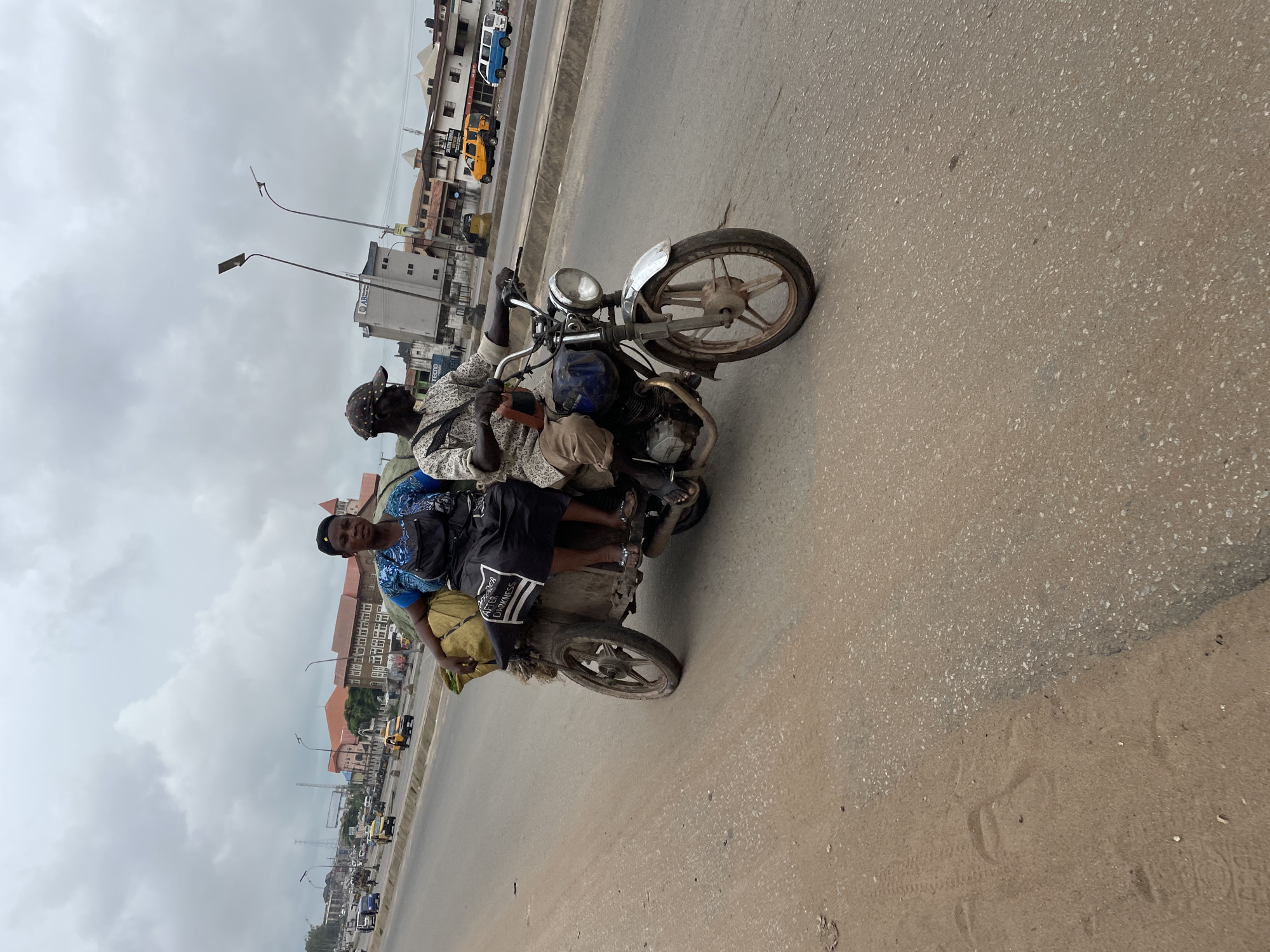 Two people, a man and a woman, are riding on a motorcycle with a sidecar. The man is driving the motorcycle, and the woman is seated in the sidecar, which is loaded with bags. They are on a paved road with some sand on the side, and there are buildings and traffic in the background. The sky is overcast. The motorcycle appears to be a common model used for transport, possibly in a developing country. The woman is wearing a blue top and a black apron, and the man is wearing a patterned shirt and a cap. They appear to be transporting goods or merchandise, as indicated by the bags in the sidecar. The scene suggests a busy urban environment with active transportation.