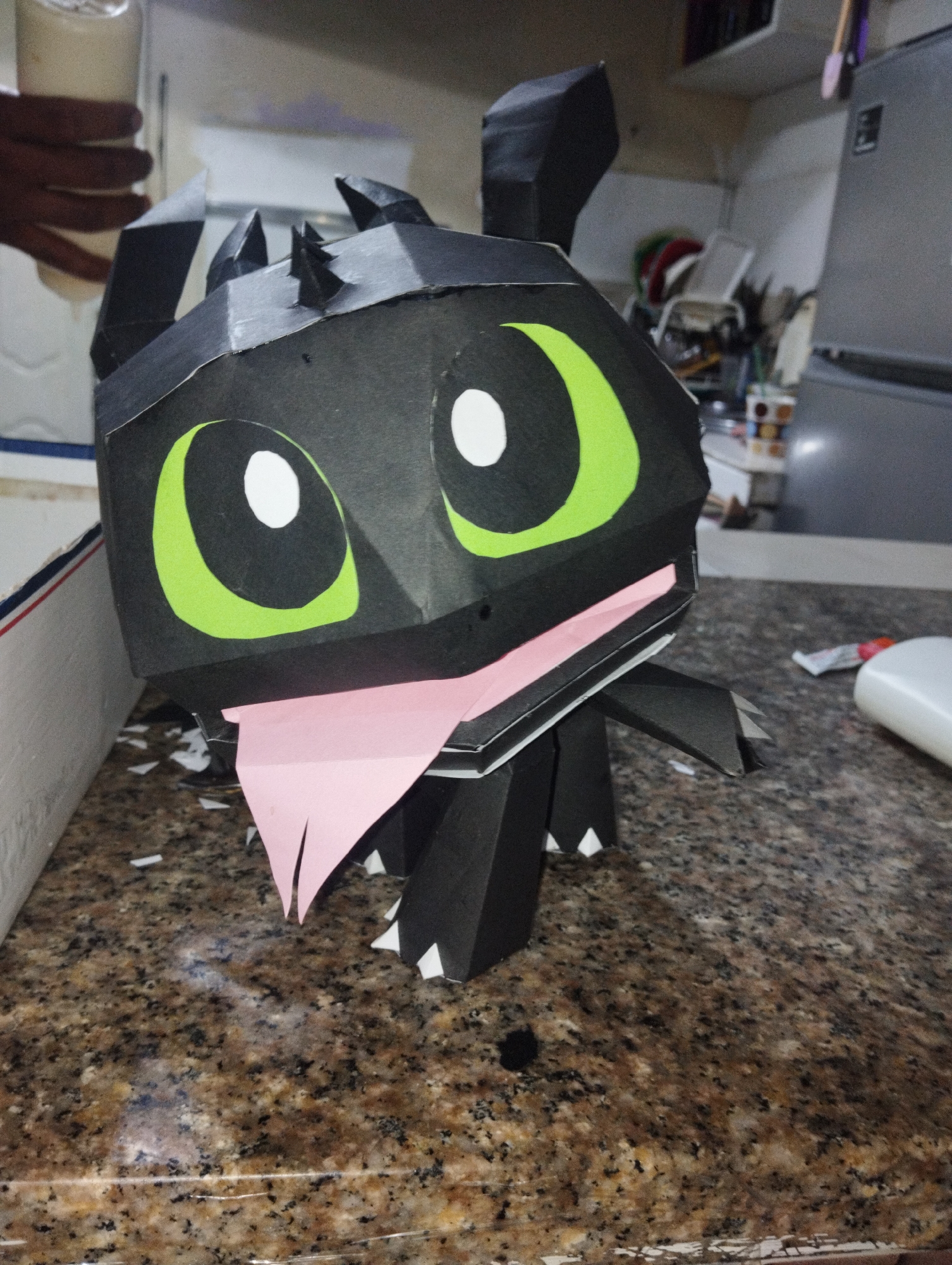 A papercraft model of Toothless from "How to Train Your Dragon" sits on a mottled brown countertop. The model is black with large green eyes and a pink tongue sticking out. The location is likely a home kitchen with white cabinets and a stainless steel refrigerator visible in the background. A person's hand holds a jar in the upper left corner. Small scraps of paper litter the countertop around the model. A white bottle is visible to the right. This scene takes place at El Valle del Espiritu Santo, Venezuela.