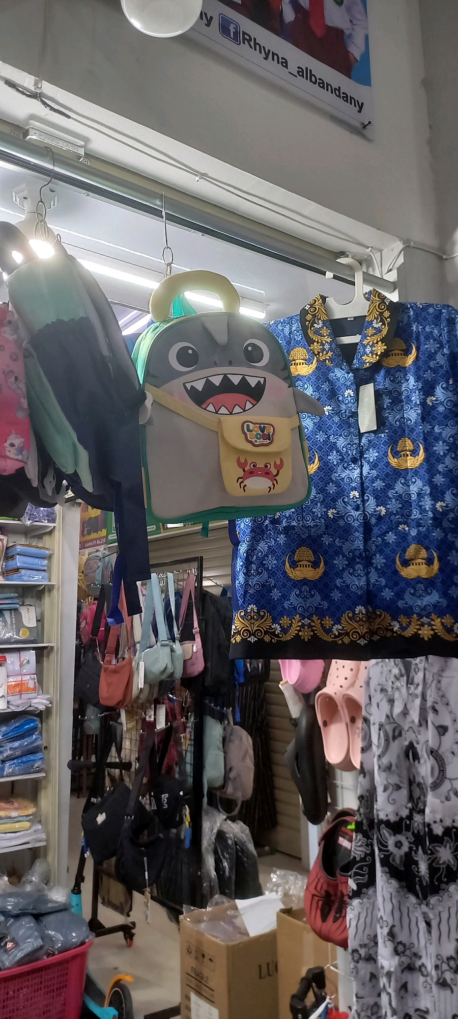 The image depicts a retail stall, likely in a marketplace, in Melayu, Indonesia. The stall displays a variety of merchandise, primarily focusing on bags and clothing. In the foreground, a child's backpack shaped like a shark with a yellow pouch featuring a crab motif is prominently displayed. To its right, a blue patterned garment, possibly a batik shirt, hangs on a hanger.
Behind these items, numerous smaller bags of various colors and styles are hung from racks. On the left side of the image, shelves are stocked with packaged goods, likely clothing or accessories, in shades of blue and white. A pink plastic basket filled with blue fabric items is visible at the bottom left.
The environment appears to be an indoor or semi-enclosed space, with a white ceiling and artificial lighting evident. A banner with text "y f Rhyna_albandany" is partially visible at the top of the image, suggesting the name of the business or vendor. The overall impression is of a vibrant, albeit cluttered, retail setting. There are no people actively engaged in the scene, but the presence of merchandise implies a commercial transaction is the purpose of the location.