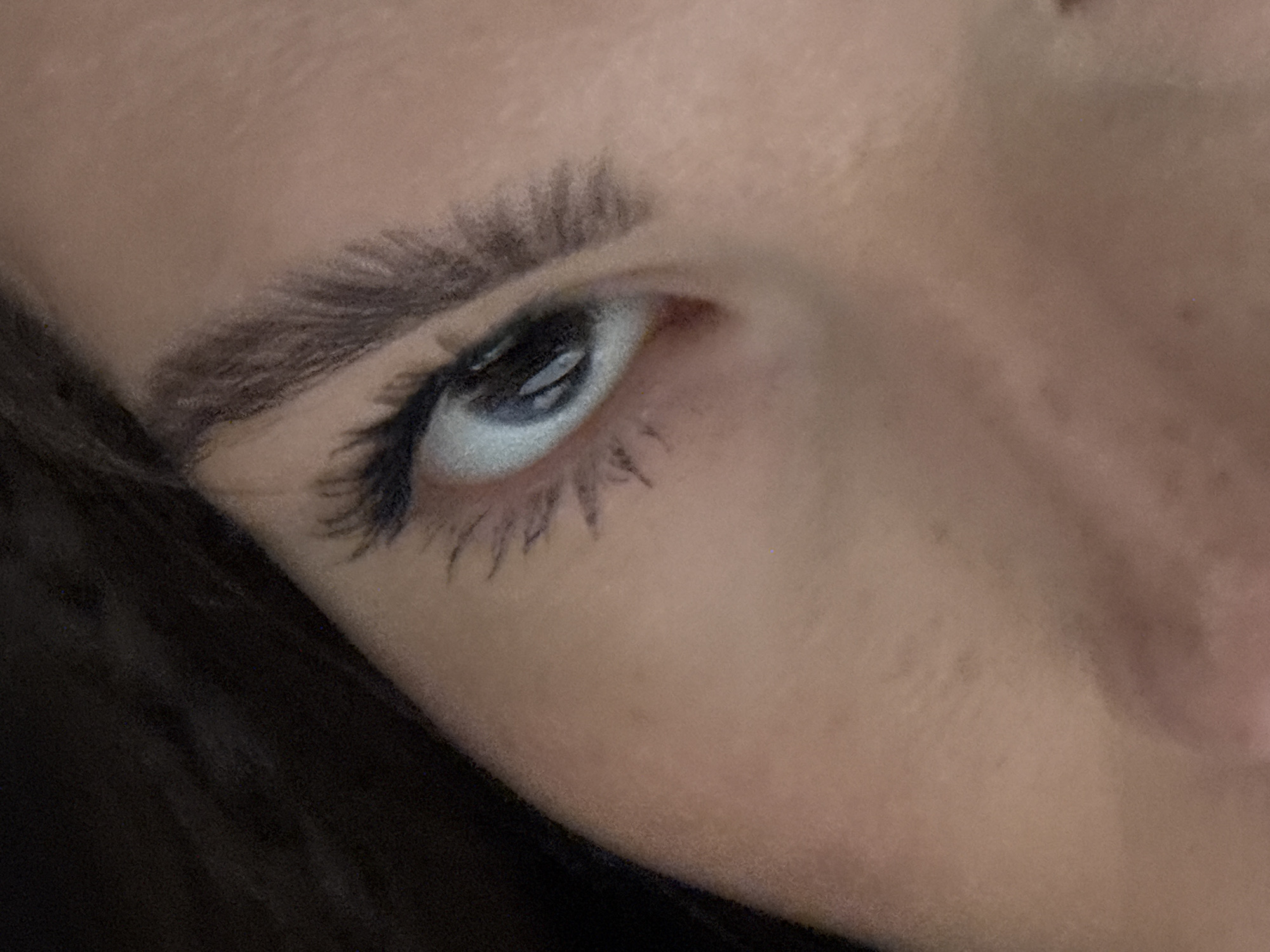A close-up, angled photograph depicts a person's left eye and eyebrow. The eyebrow is dark and thick. The eye is partially open, with visible upper and lower eyelashes coated in mascara. The skin tone is fair. The angle of the photograph reveals part of the person's face, including the nose and hair. The scene takes place indoors with neutral lighting. No specific location in Шемордан, Russia is identifiable.