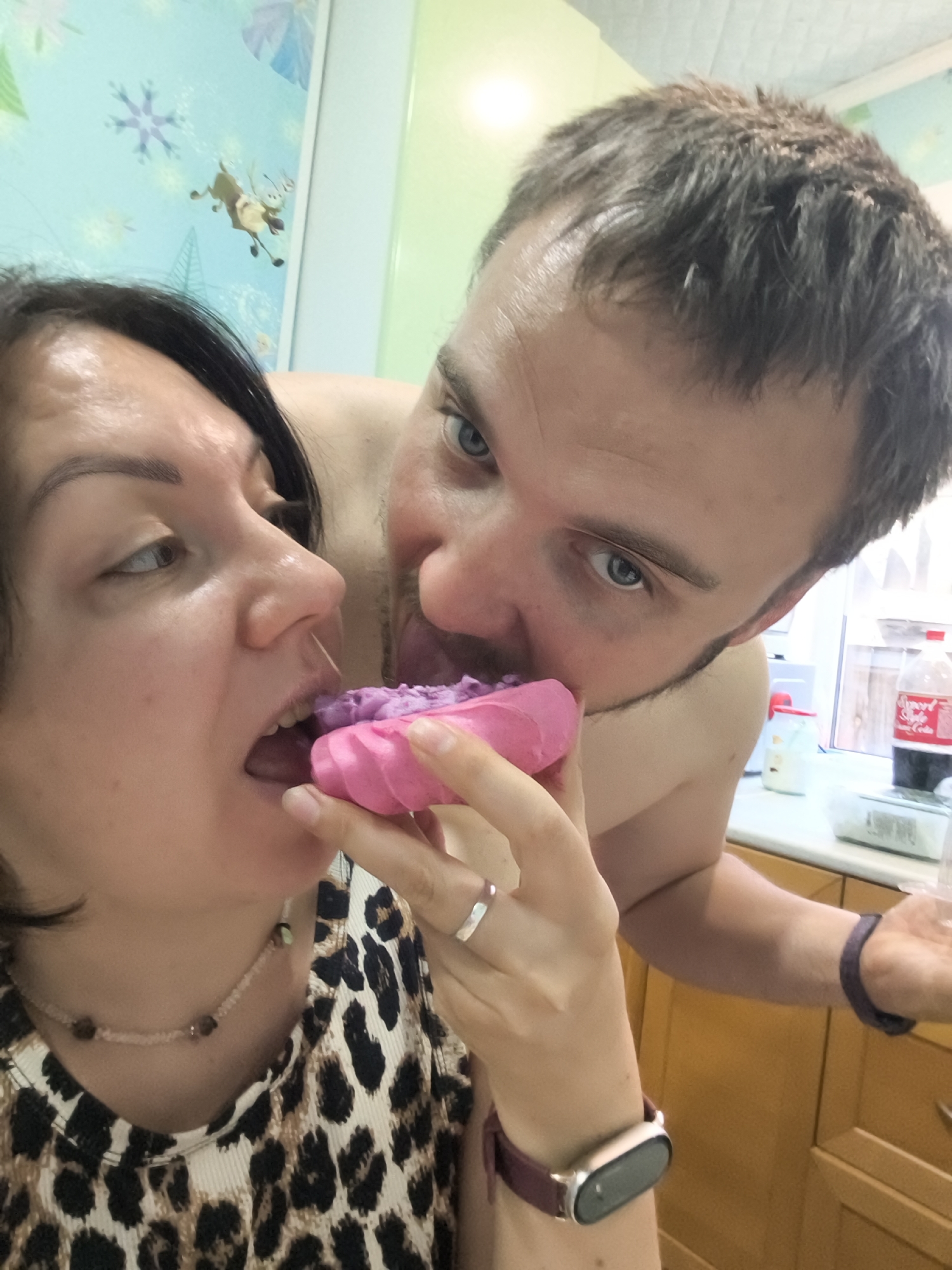 A medium shot captures a close-up of a woman and a man engaged in a playful interaction. The woman, positioned on the left, has dark hair and is looking towards the left side of the frame with her mouth open, in the process of taking a bite from a pink pastry. The man, on the right, is leaning towards her, his mouth open and his tongue extended towards the pastry, seemingly trying to share the bite. Both individuals appear to be in a domestic setting, possibly a kitchen, with wooden cabinets visible in the background. The lighting is bright, suggesting an indoor environment. The image is captured from a slightly low angle, looking up at the subjects.