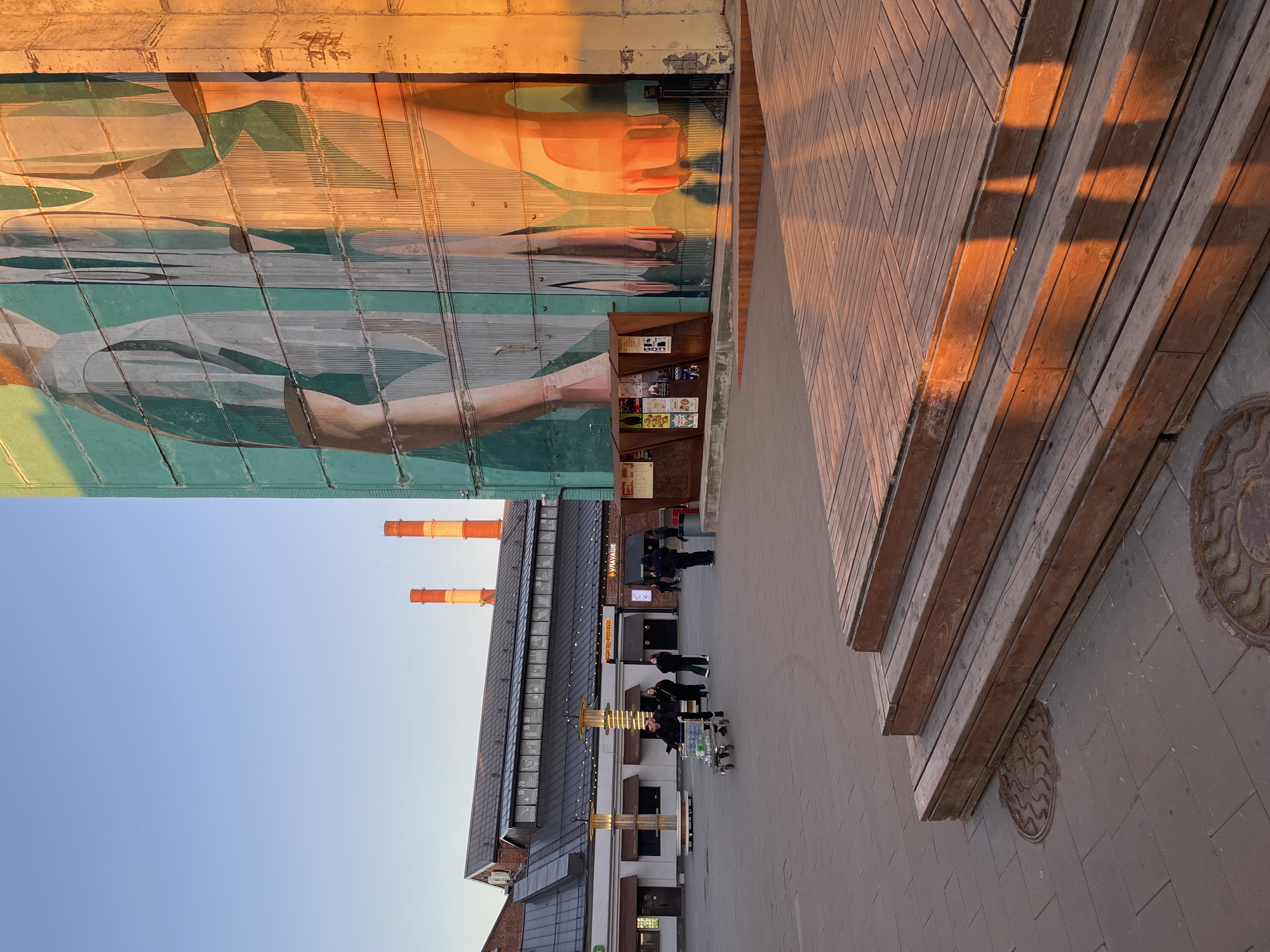 The image depicts a scene in Saint Petersburg, Russia. It features a large mural of a human hand on a building facade, accompanied by a small bookshelf. Adjacent to the mural is a wooden deck with steps. The ground is paved with grey stone tiles. In the background, a building with red and white smokestacks is visible against a clear blue sky. Several individuals are walking along the paved area, and a shopping cart is also visible.