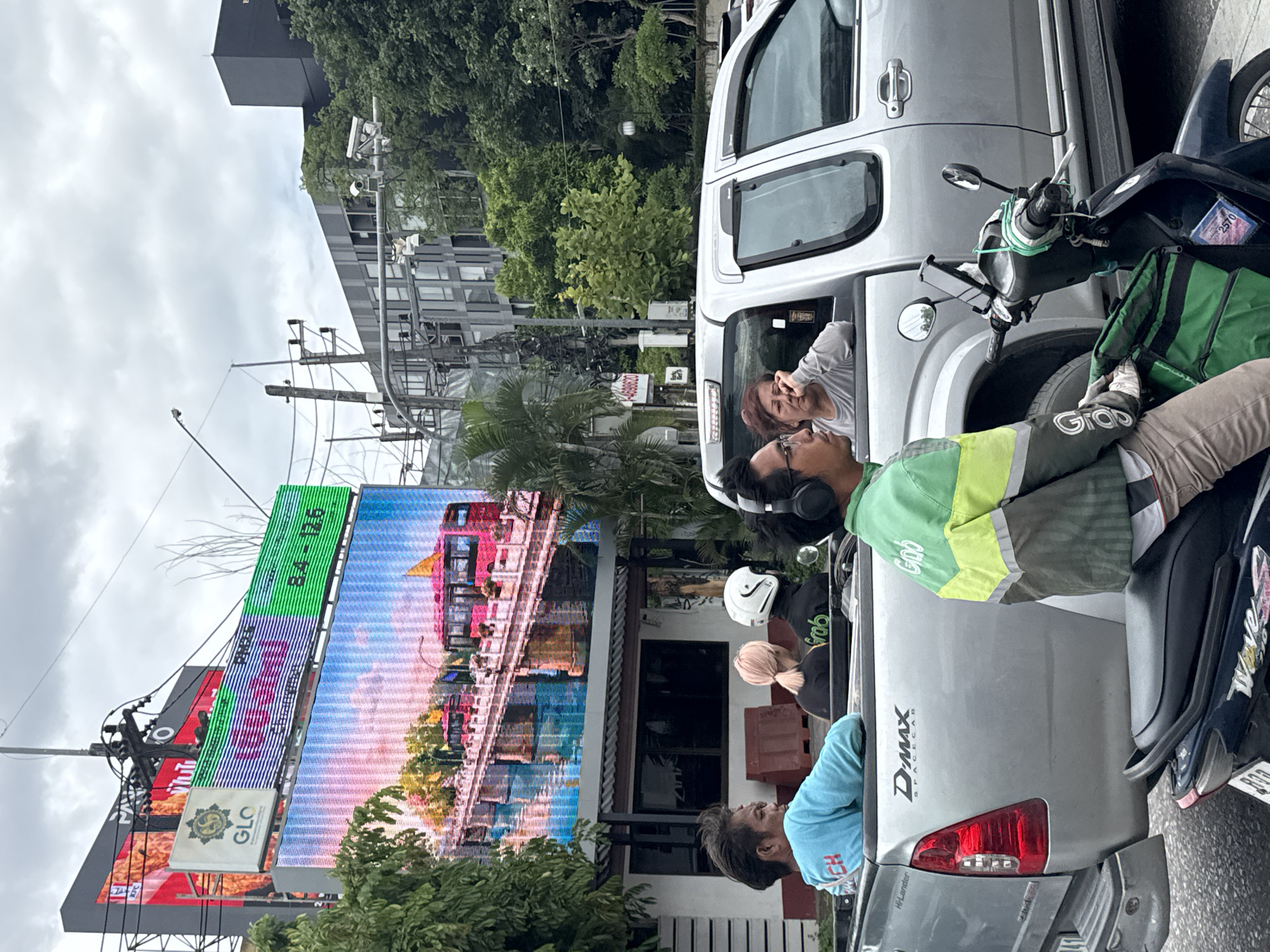 In Chiang Mai City Municipality, Thailand, on a cloudy day, a silver pickup truck with a D-MAX Spacecab designation is stopped in traffic. In the truck bed, four individuals are seated: a man in a blue t-shirt with "CH" written on it, a woman with light-colored hair, a person wearing a white motorcycle helmet, and a woman in a grey shirt looking away. Ahead of the pickup truck, a person wearing a green and yellow reflective jacket for "Grab" is seated on a dark-colored motorcycle. This person is wearing headphones and appears to be looking forward. The background features a large digital billboard displaying a colorful image of a bridge and a red bus, alongside a sign with "PM2.5" and numerical data. Above the billboard, other signs are visible, including one for "GLO" and a red KFC sign. Buildings and trees line the street, with power lines and streetlights also present. The scene depicts a typical urban traffic situation with various modes of transportation and advertising.