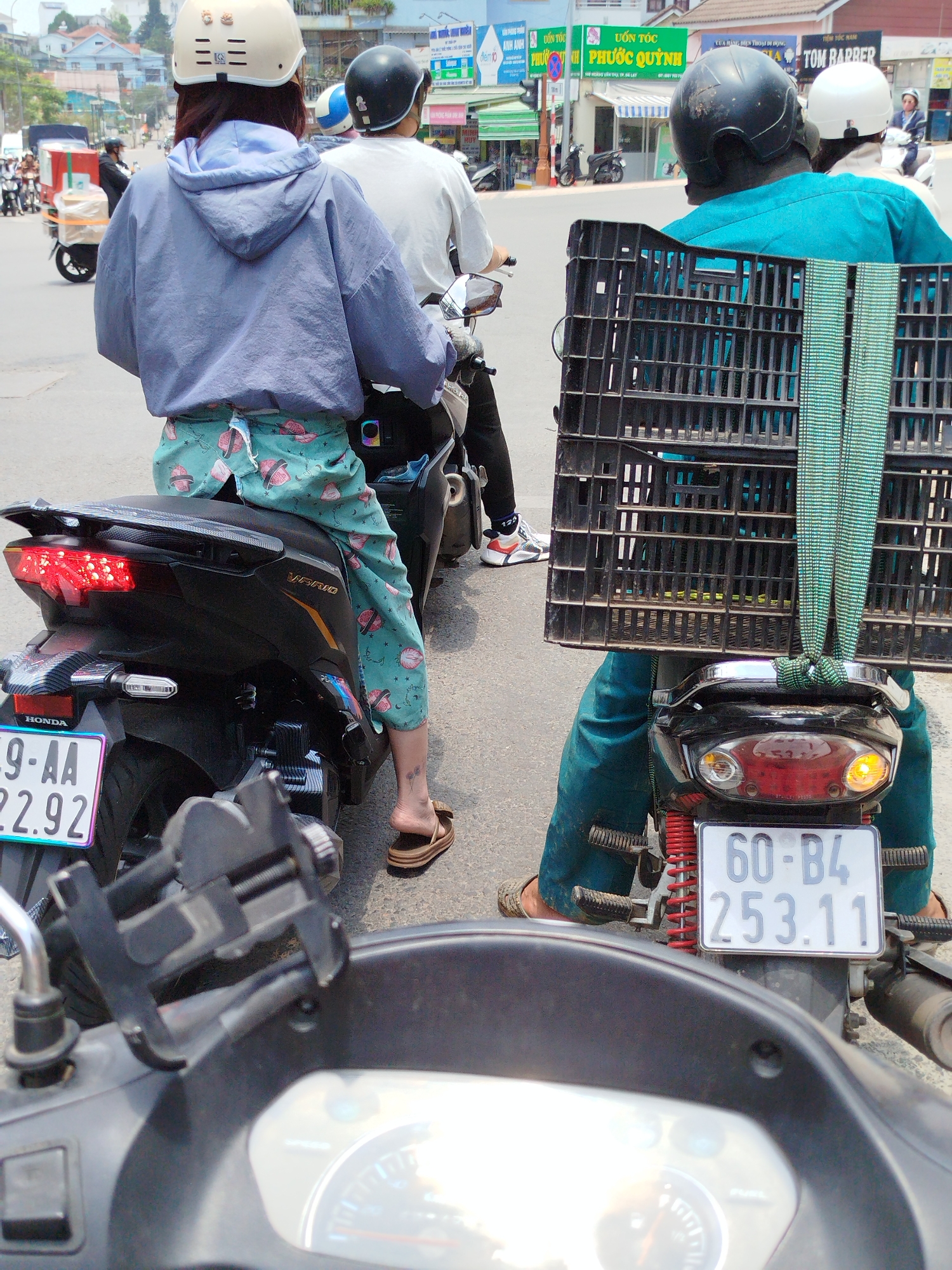 The image is a street scene in Da Lat, Vietnam, showing a line of motorbikes at a standstill. The focal point is the rear view of three motorbikes and their riders. The first motorbike, closest to the viewer, has a license plate reading "69-AA 22.92." The rider in front is wearing a light blue hoodie and patterned teal bottoms. The second rider is wearing a white shirt, and the third rider has a large black plastic crate strapped to the back of their scooter. This rider is wearing a blue shirt. The second motorcycle license plate reads "60-B4 253.11." The riders are all wearing helmets. In the background, there are various shops with Vietnamese signage, including a barber shop named "TOM BARBER" and a sign that includes "PHƯỚC QUỲNH". Other motorbikes and pedestrians are visible in the distance.