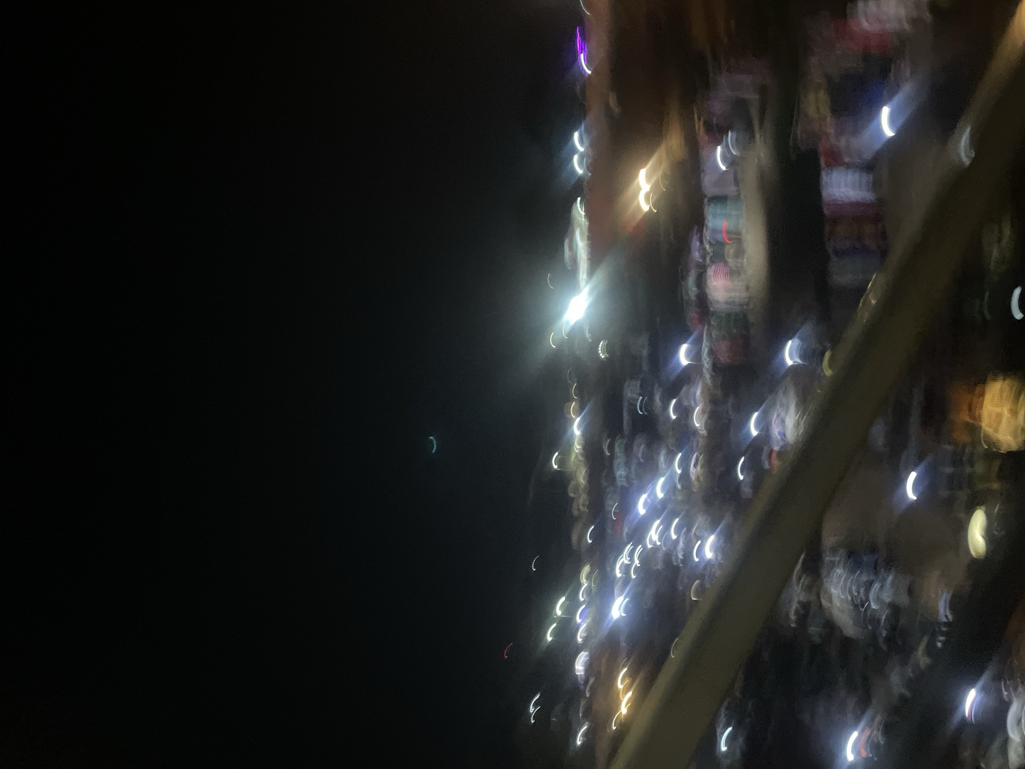 The image depicts a night scene in Sauka, Nigeria, characterized by blurred lights and indistinct forms, suggesting motion blur or a low-light setting. The foreground features a dark, sloping structure, possibly a railing or barrier. Beyond this, a multitude of colorful, elongated light streaks, some appearing as crescents, are scattered across the scene, indicating a busy urban environment with moving lights, such as vehicles or streetlights. Some areas show clusters of what appear to be stalls or displays with varying colors, hinting at a marketplace or commercial area at night. A faint, pale green arc is visible in the upper portion of the sky, which could be a sliver of the moon or atmospheric light. The overall impression is one of a vibrant, active night in the city, albeit rendered abstractly due to the photographic effects.