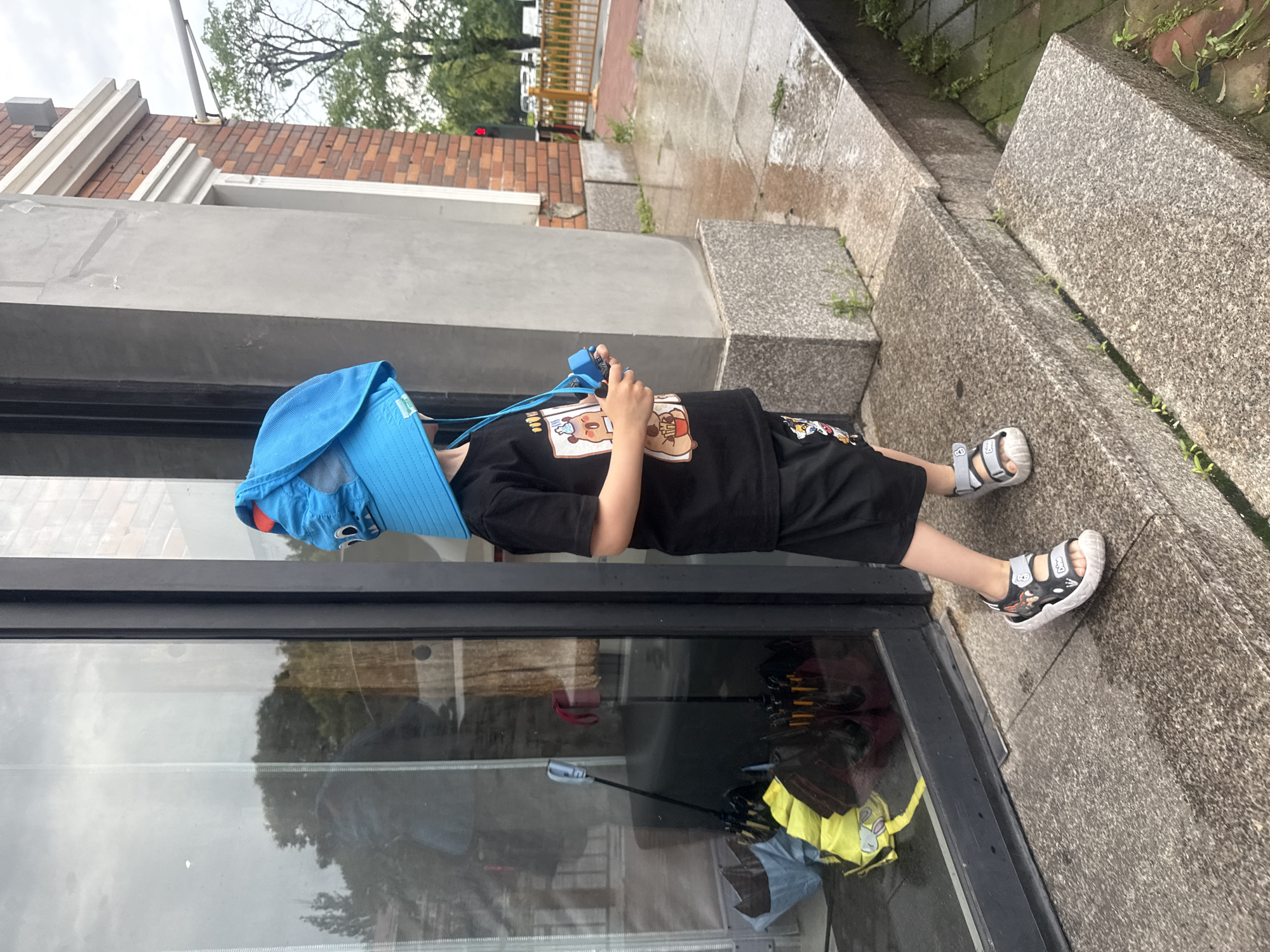 A young child stands on a stone staircase outside a building, wearing a blue hat with ear-like flaps, a black t-shirt with a graphic of a cartoon bear, and black shorts with a similar graphic. The child is holding a blue toy in their right hand. The ground appears wet, suggesting recent rain. In the background, a large glass window reflects trees and the sky. Several umbrellas are visible inside the building through the window. The architecture suggests an urban setting, possibly in Qinhuangdao, China, given the presence of red brickwork and a traffic light visible further down the street. The scene appears to be a casual moment captured outdoors.