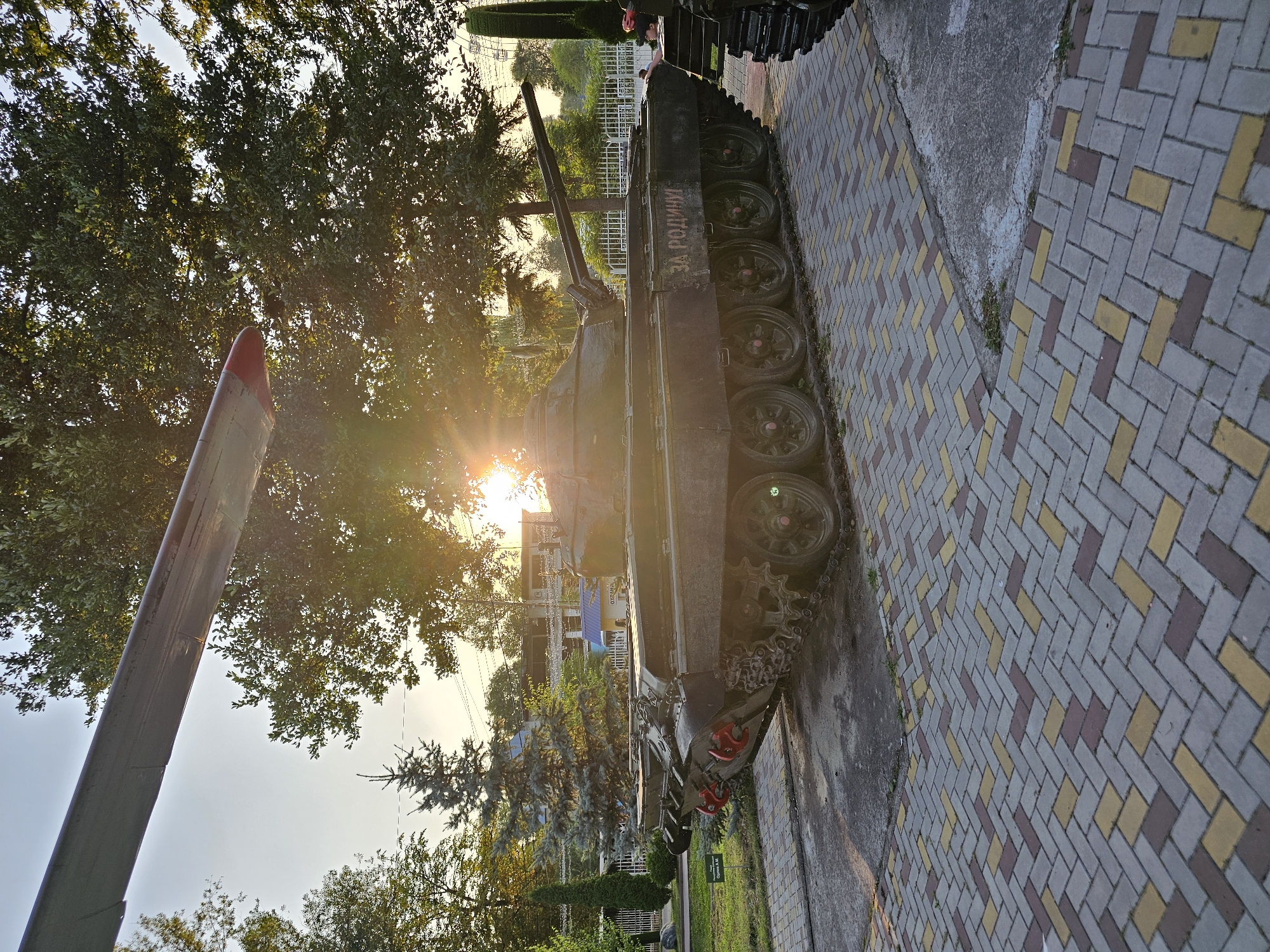 An old tank, marked with the Cyrillic inscription "ЗА РОДИНУ" (For the Motherland), is displayed outdoors on a paved area in Cherkessk, Russia. The tank, which appears to be a Soviet-era model, is positioned on a herringbone-patterned brick walkway. Behind the tank, trees create a backdrop with sunlight filtering through their leaves, suggesting it is late afternoon or early evening. An airplane wing, detached and seemingly part of a display, juts into the frame from the upper left corner. In the distance, a Ferris wheel is visible, hinting at a possible amusement park or memorial complex. A person can be seen near the rear of the tank, interacting with it. The overall scene is an outdoor exhibition featuring military hardware and possibly located in a public space within the city of Cherkessk.