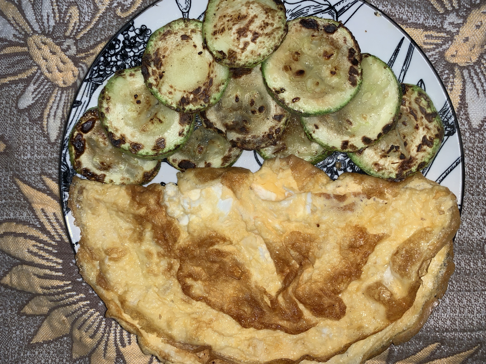 An overhead shot captures a plate of cooked food resting on a patterned tablecloth. The meal consists of two distinct components: a pile of pan-fried or grilled zucchini slices and a large, folded omelette or fried eggs.
The zucchini slices are arranged in the upper half of a white plate, which features a decorative black and white intricate pattern along its rim. The slices are round, varying in color from light green to a pale yellow, and exhibit noticeable browning and charring in various spots, indicative of having been cooked on a hot surface. Some slices appear slightly translucent.
Occupying the lower portion of the plate is a substantial serving of eggs, appearing as a thick, folded omelette or a large mass of fried eggs. Its surface displays a range of golden-yellow to deep brown colors, with some areas more caramelized and crisped than others. Lighter, creamy white sections of cooked egg are also visible within the folds.
The plate is placed on a textured fabric surface, likely a tablecloth, which is adorned with a prominent floral or leaf-like pattern in shades of brown, beige, and gold. The overall lighting suggests an indoor setting. There are no people, specific location markers, or visible text in the image. The combination of eggs and vegetables implies a breakfast, brunch, or light meal.