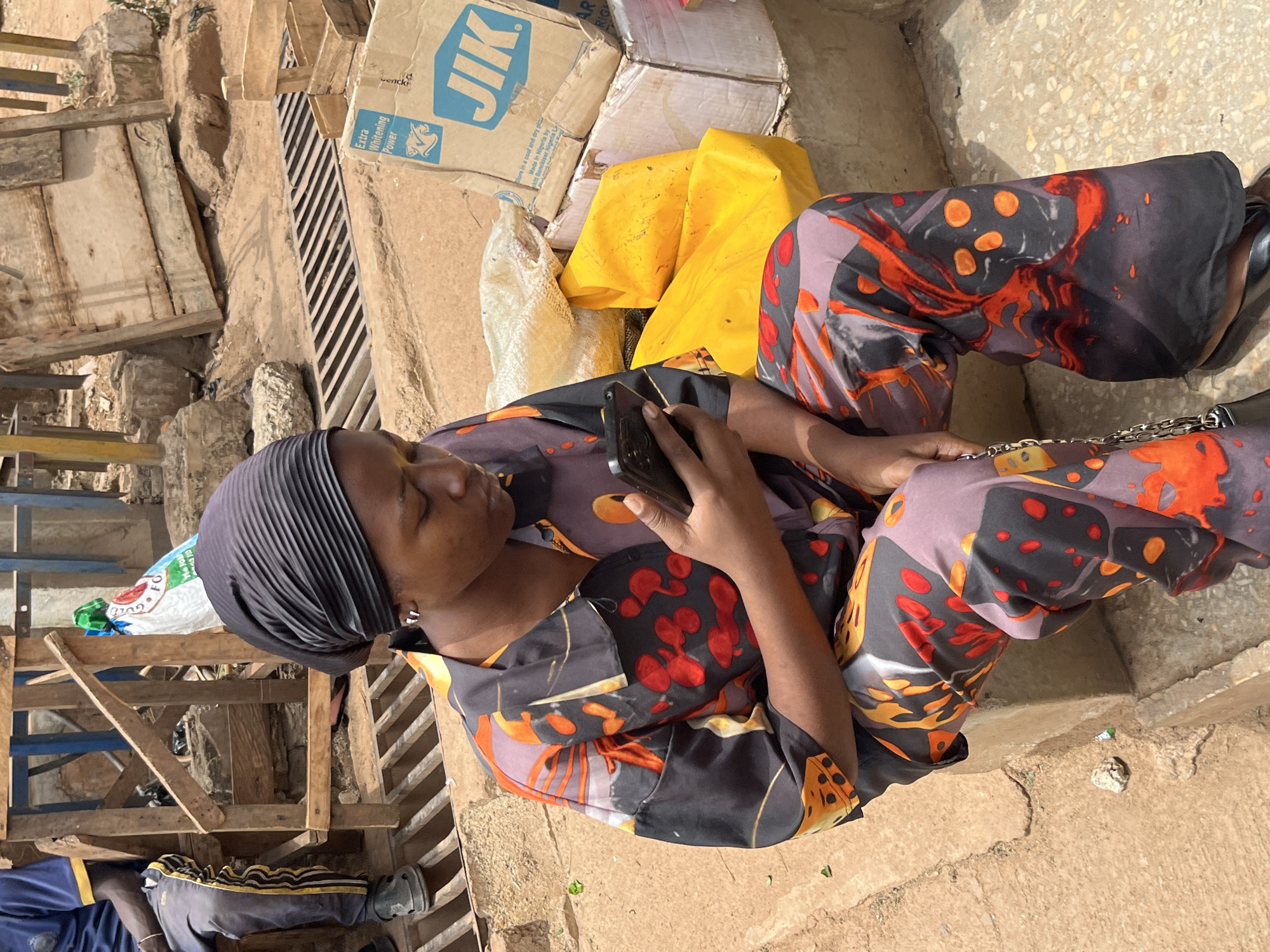 A woman in a headscarf and patterned clothing sits on a concrete surface, possibly a step or ledge, in what appears to be an outdoor marketplace or urban area in Jos, Nigeria. She is holding a smartphone and looking at it. Beside her are various objects including cardboard boxes labeled "JIK" and some bundled items in yellow and white cloth. The ground is concrete and there is a wooden structure and a metal grate in the background.