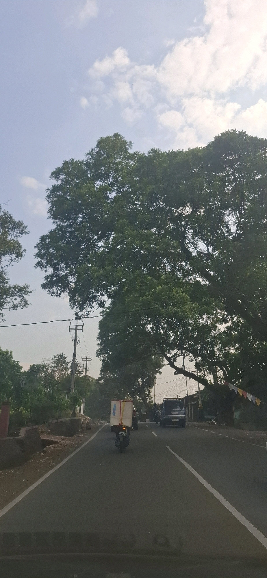 The image depicts a street scene in Cilaku, Indonesia, under a cloudy sky. A large tree with dense green foliage dominates the right side of the frame, its branches extending over the road. The road is paved and has a white line marking the center. In the foreground, the edge of a vehicle's dashboard is visible. On the road, a motorcycle with a large white box secured to its rear is traveling away from the viewer. A dark-colored van is also on the road, further ahead and to the right of the motorcycle. On the left side of the road, there are some rocks and vegetation, and a utility pole with electrical wires. The overall impression is of a quiet, slightly overcast day on a road lined with trees.