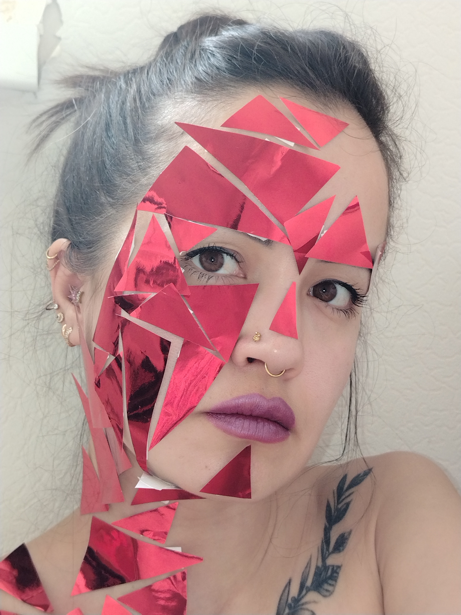 A close-up studio shot depicts a woman with her hair pulled back into a bun, adorned with reflective red triangular pieces partially covering her face and chest. She has brown eyes, a gold nose ring and stud, and wears mauve lipstick. A tattoo of a leafy plant is visible on her shoulder. The background is a plain white wall. The location is not provided in the image.