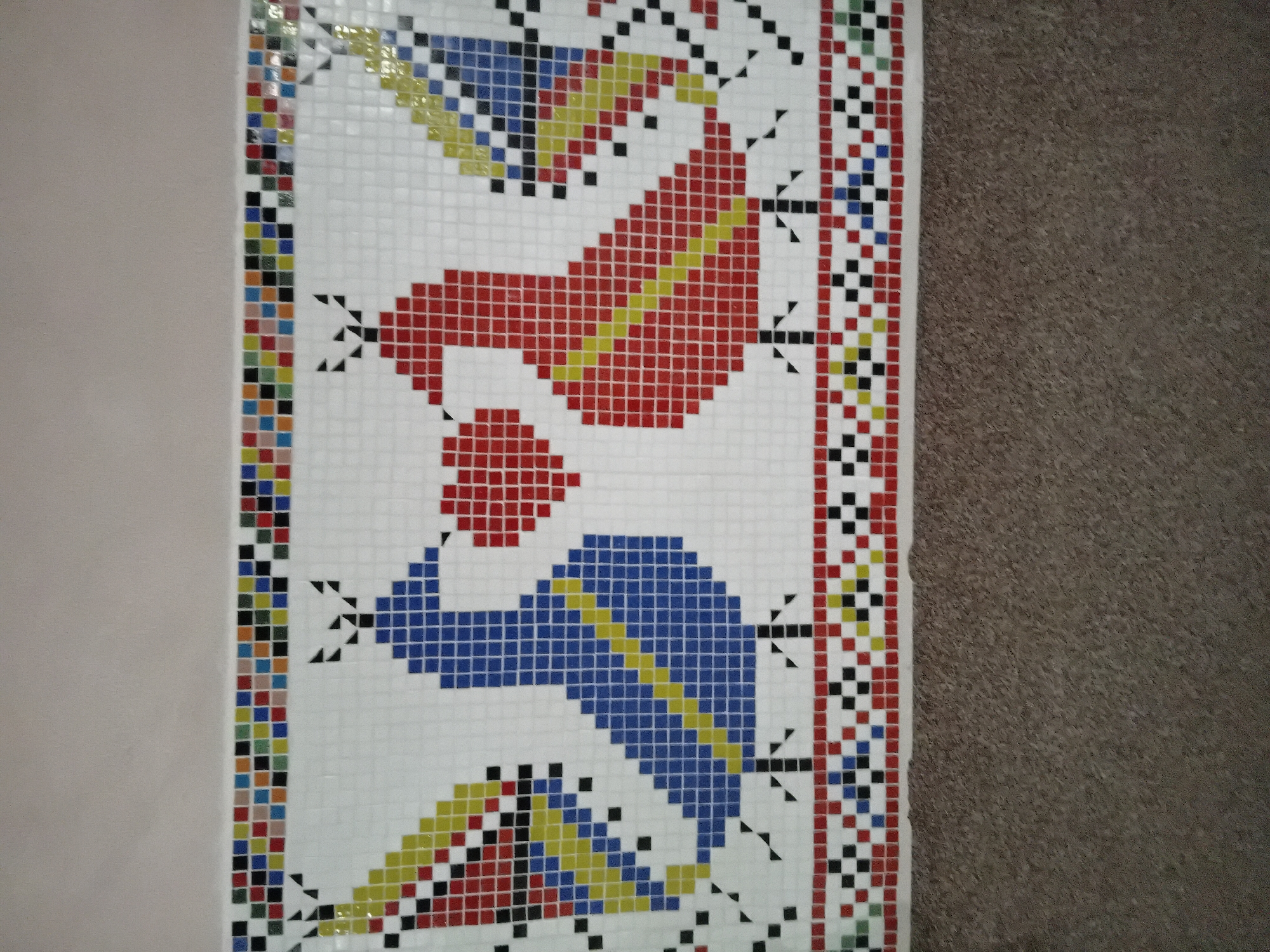 An overhead view captures a vibrant mosaic artwork featuring stylized figures and geometric patterns. The mosaic is composed of numerous small, square tiles set against a white tile background.
The central design showcases two large, abstract figures in profile, facing each other. The upper figure is predominantly red with a diagonal yellow stripe, while the lower figure is primarily blue, also with a diagonal yellow stripe. Both figures have elements suggestive of heads or headdresses, depicted in yellow and blue, respectively. Around these main figures are smaller, abstract shapes and lines in black, resembling arrows or decorative flourishes.
The entire mosaic is framed by a colorful, multi-tile border that runs vertically along both sides of the central design. This border consists of a repeating pattern of small red, blue, green, yellow, black, and white squares, creating a lively contrast.
To the left of the mosaic, a plain, light-colored wall or surface is visible, providing a neutral backdrop. To the right, a darker, textured surface, possibly a carpet or another type of flooring, fills the remainder of the frame. The image is taken from a slightly elevated and angled perspective, giving a dynamic view of the tile work. There is no visible text.