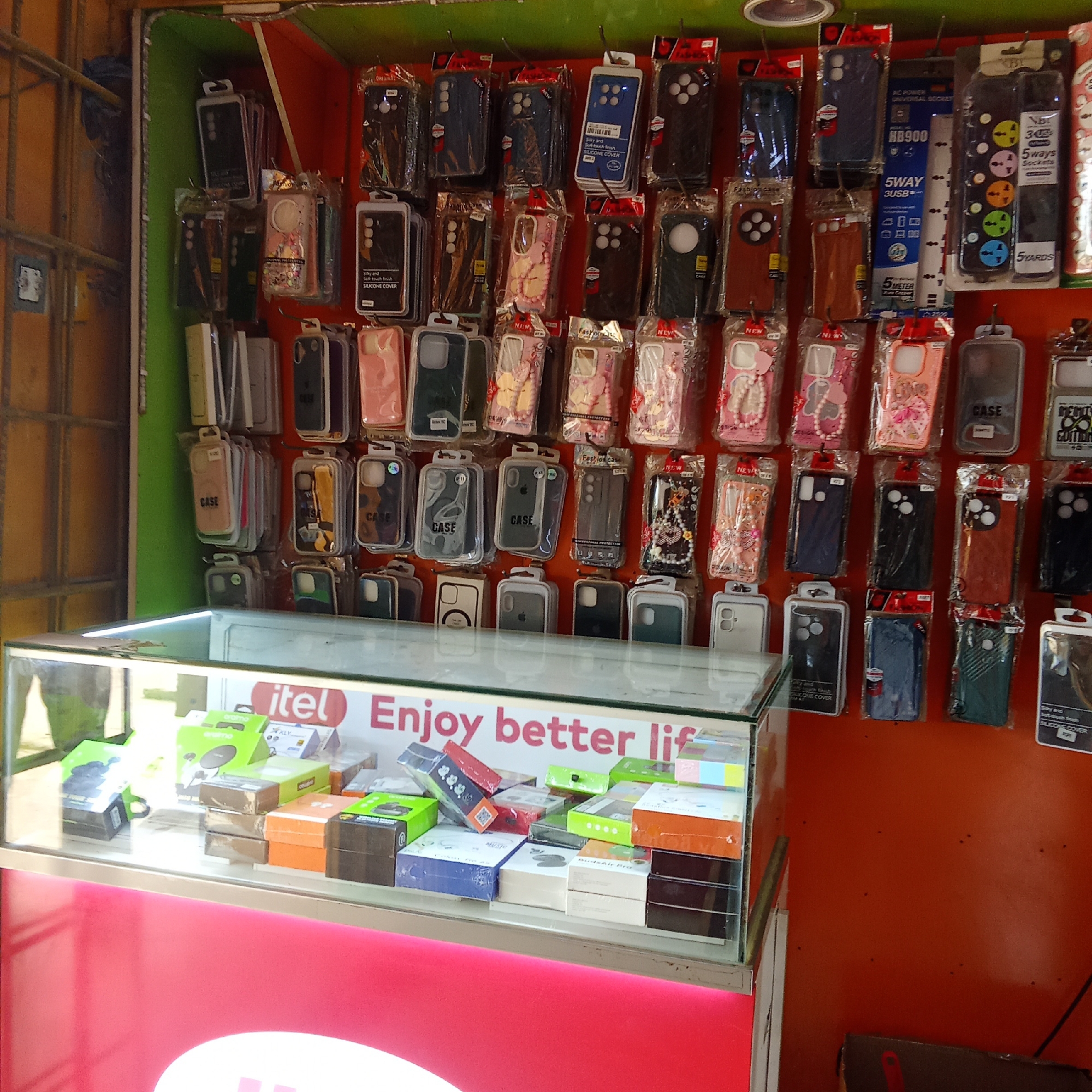 The image depicts a small retail shop specializing in mobile phone accessories. Displayed on the wall are numerous mobile phone cases of various styles and colors, still packaged. A glass display case below showcases boxed accessories, including earphones and chargers. The "itel" brand logo is prominently featured on the case, along with the slogan "Enjoy better lif." The overall setting suggests a street-side vendor or a small shop located in an urban environment.