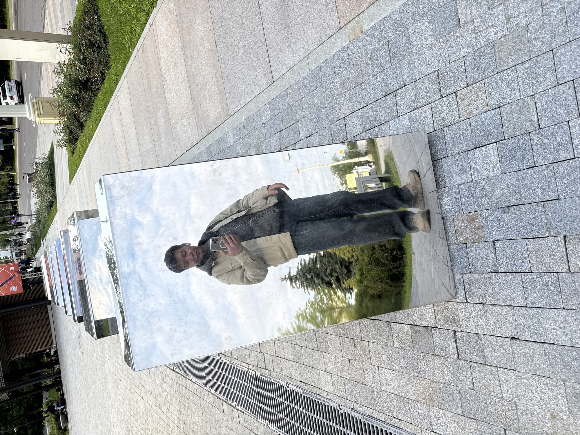 The image, rotated 90 degrees counter-clockwise, depicts a man reflected in one of several large, rectangular mirrored panels that form a public art installation. The man is the primary subject, shown from the waist up, wearing a light beige jacket over a dark garment, dark pants, and light-colored shoes. He is holding a silver smartphone up to take a picture or selfie of his reflection. His face, reflected in the mirror, shows him with short dark hair, looking towards his phone, and appearing to smile slightly.
The mirrored surface reflects a bright, slightly cloudy sky, lush green trees, and bushes. The ground around the installation is paved with light grey, textured stone blocks, likely granite, with a darker grey section adjacent to the mirrors and a lighter, smoother path further away. A long metal drainage grate with horizontal bars is visible in the immediate foreground, running parallel to the art installation.
In the background, visible both directly and through reflections, are more green foliage, a road, and architectural elements including a white column and a white SUV (possibly a BMW X3). Other mirrored panels extend into the distance, reflecting more of the urban environment. The setting appears to be a public park or plaza in Moscow, Russia, on a clear day, likely late morning or afternoon given the bright but not harsh lighting.
Visible text on the drainage grate includes "Л-500", "ДСТУ Б В.2.5-26:2005", and "400X500ММ", which are technical specifications or a product standard. A distant red and white sign with graphics is indistinctly reflected on one of the other panels.