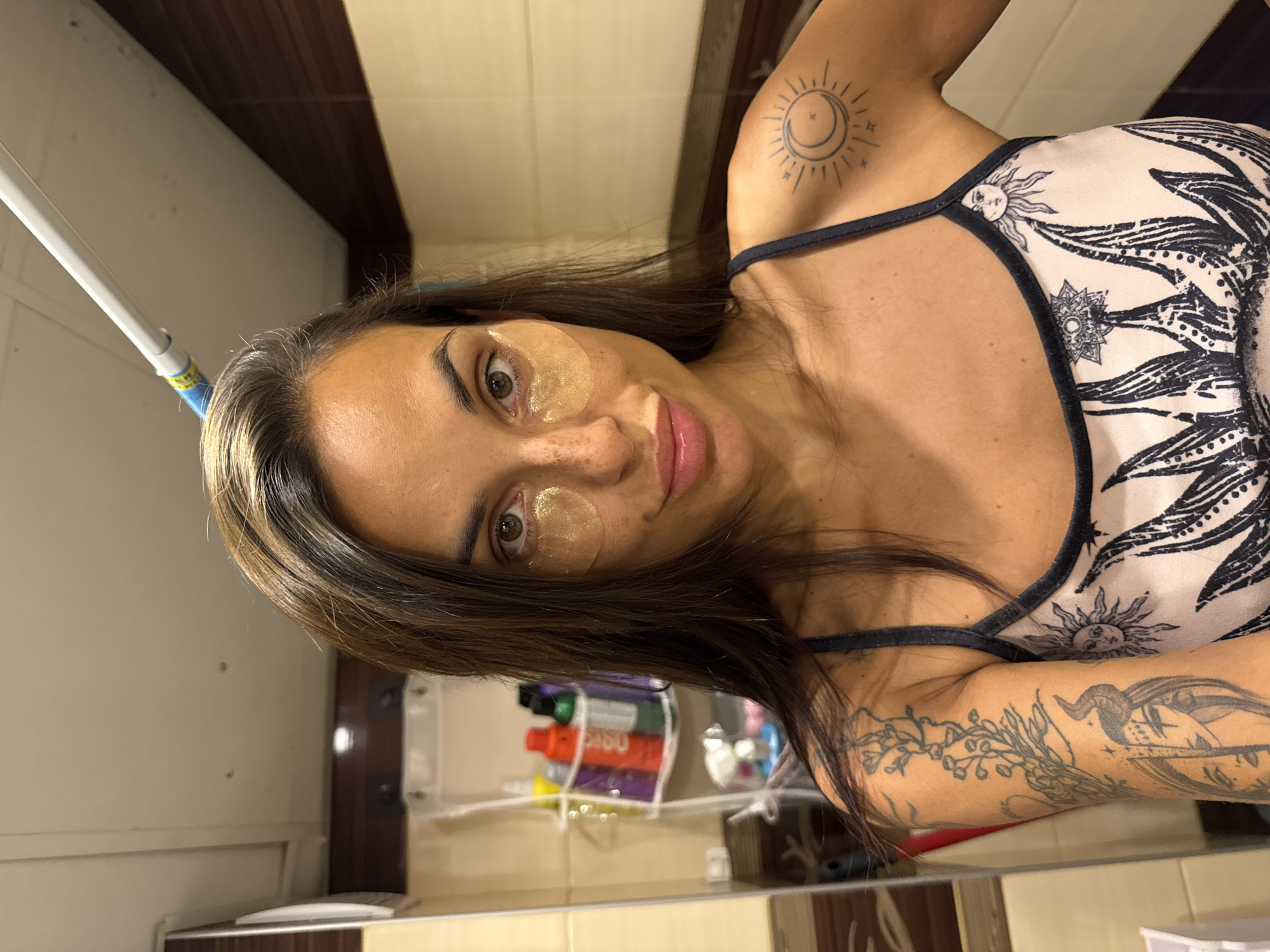 The image shows a woman with dark hair and tan skin in what appears to be a bathroom. She is wearing gold under-eye patches and a camisole with a sun and moon print. She has tattoos on her arm and shoulder, including a sun and moon design on her shoulder. In the background, a shelf is visible with various bottles and containers, possibly toiletries. The environment suggests an indoor setting, likely a residential bathroom. There is no information to suggest the image is located in Pleven, Bulgaria.
