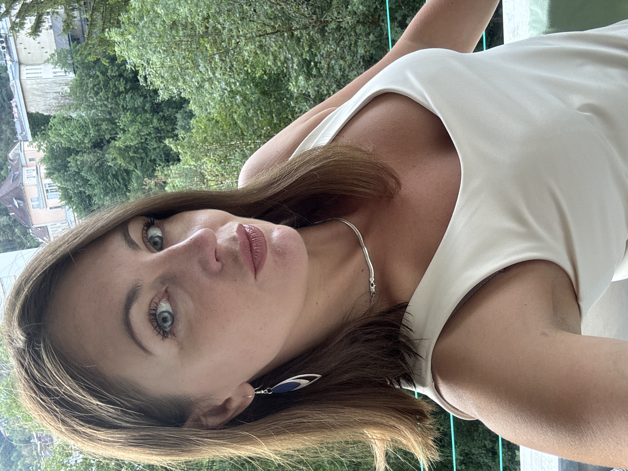A woman with shoulder-length brown hair and blue eyes is taking a selfie. She is wearing a light-colored tank top, a silver necklace, and a long, dark blue and silver earring in her left ear. Her skin appears lightly tanned, and she has a subtle makeup application. She is indoors, possibly on a balcony, with a lush green forest and buildings visible in the background. The architecture in the background suggests a European setting, consistent with the city of Saint-Claude, France, known for its picturesque natural surroundings and historical buildings. The lighting indicates it is daytime.