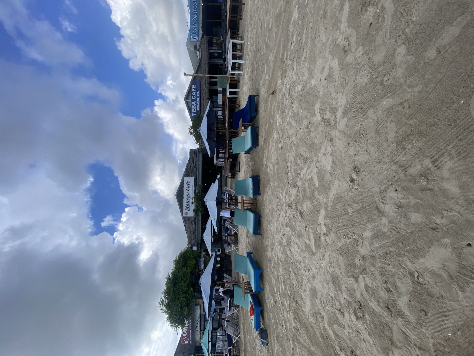 The image shows a beach scene in Kuta Selatan, Indonesia, with several beach chairs and umbrellas set up on the sand. In the background, there are a few cafes and restaurants, including "Menega Cafe" and "TEBA CAFE JIMBARAN - BALI". The sky is partly cloudy with patches of blue sky visible. The sand is light brown and appears to be packed down in some areas, with some tire tracks or footprints visible. There are several lounge chairs, some with blue cushions, arranged in rows. Umbrellas are also present, providing shade over some of the seating areas. A few people are visible in the distance, near the buildings, possibly staff members at the cafes. The overall impression is a relaxed beachside dining or lounging area.