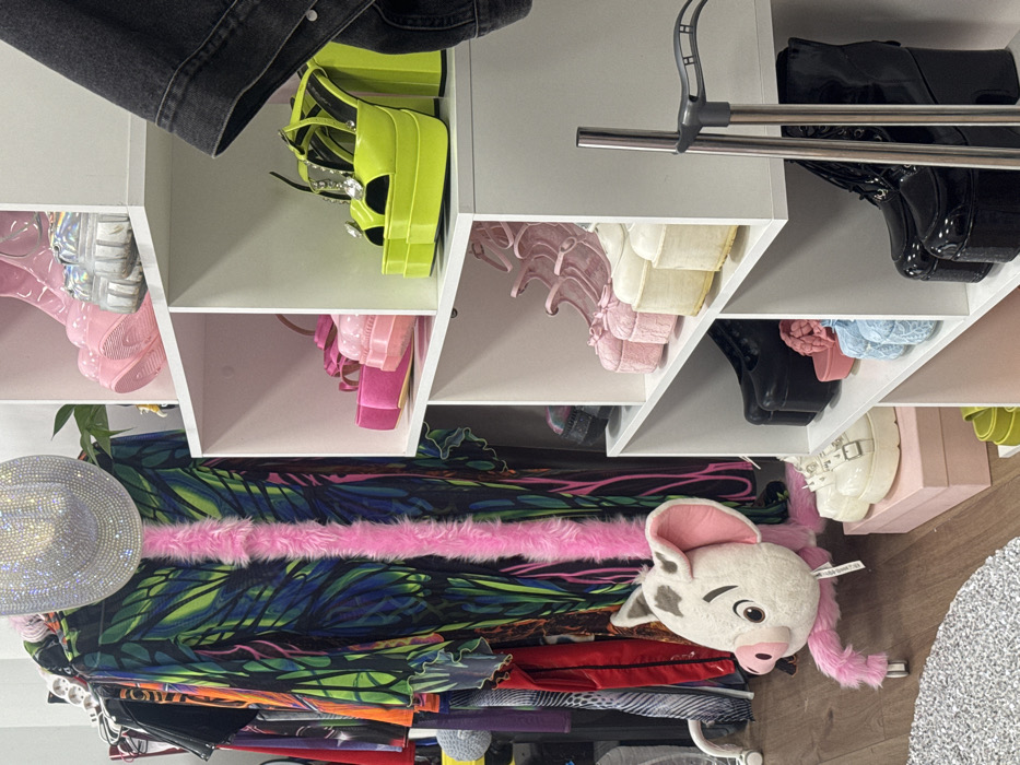 This image displays the interior of a commercial space, likely a retail boutique, within the city of Sofia, Bulgaria. The specific address or landmark is not identifiable from the visual evidence.
The scene features a diverse collection of fashion items, primarily clothing and footwear, arranged for display. On the left, a clothing rack holds multiple garments, including a prominent long garment with a vibrant green, blue, and black tropical leaf pattern, centrally adorned with a vertical strip of bright pink faux fur. A sparkling, rhinestone-covered cowboy hat is positioned on top of this garment. Below the clothing rack, on the light wood-toned floor, a large plush toy resembling the pig character Pua from Disney's "Moana" is present, equipped with pink furry legs on small wheels. A portion of a textured, light grey rug is visible in the lower-left foreground.
The right side of the image is occupied by a white cube-style shelving unit showcasing various high-platform women's shoes. From top to bottom, visible footwear includes: pink platform boots and silver holographic platform sneakers; bright neon yellow open-toe platform heels with crystal embellishments alongside pink platform sandals; multiple pairs of pastel-colored (pink, cream, light blue) platform Mary Jane-style shoes, some with textured details; and black platform Mary Janes, white platform combat boots with buckle straps (resting on a pink box), and black platform lace-up boots. A dark denim jacket hangs partially into the frame from the upper right, and a vertical metal pole, consistent with a display fixture, is visible on the right side. No individuals are present in the scene.