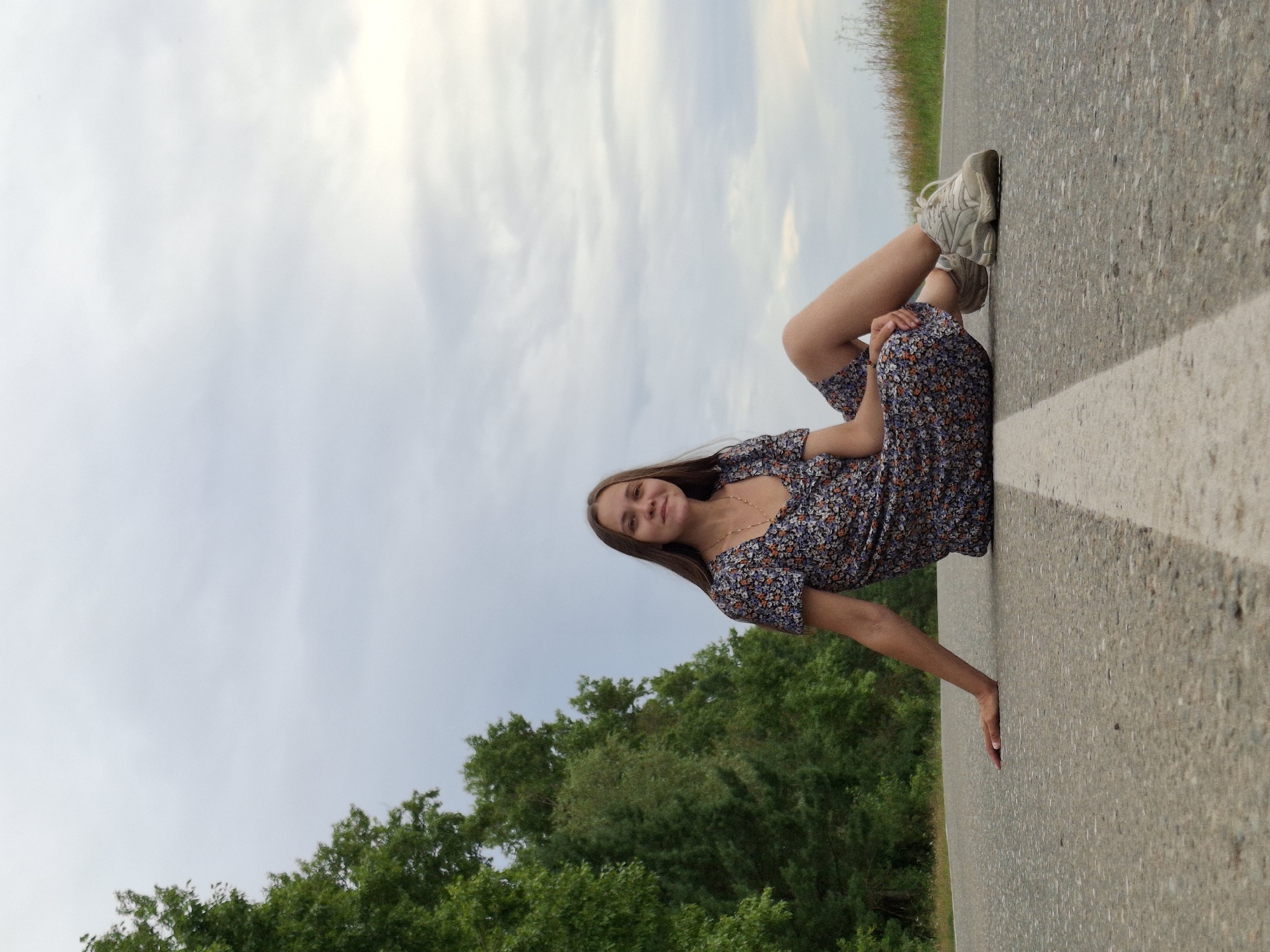 A young woman with long brown hair is sitting on an asphalt road in a rural area, possibly near the city of Garevaya, Russia. She is wearing a floral-patterned dress and white athletic shoes. Her left arm is resting on the road behind her, and her legs are extended forward. The road has a white line painted down the center. To the left of the road is a dense line of green trees, and to the right, the landscape appears to be a field with dry grass. The sky above is overcast with light gray clouds. The perspective is low, with the camera positioned on the road surface, looking up at the woman.