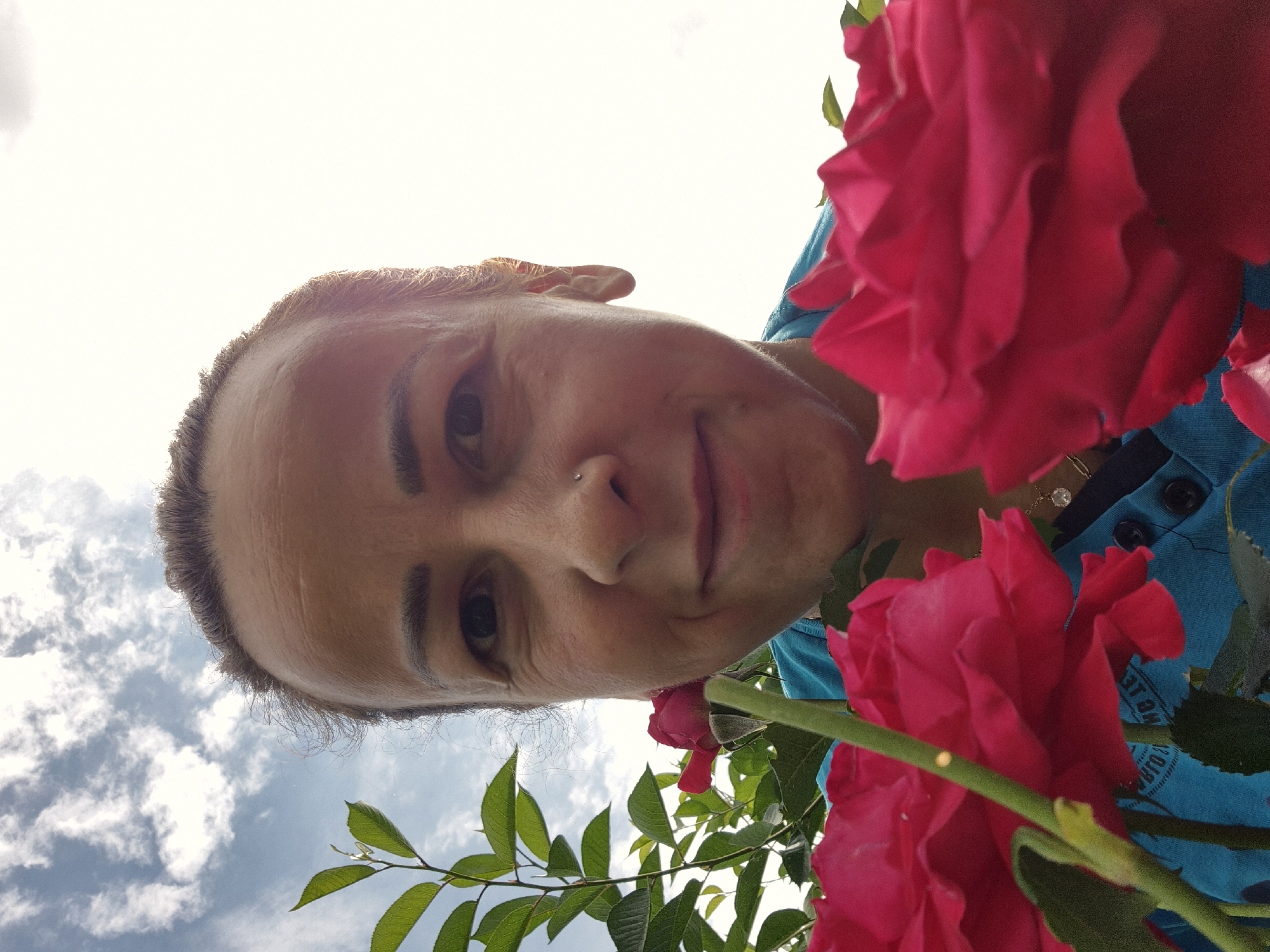 A woman is captured in a low-angle, close-up selfie, with two vibrant pink roses prominently featured in the foreground. The woman's face is angled towards the camera, and she is smiling. Her hair is pulled back, and she has a nose ring. She is wearing a blue shirt. Behind her, the sky is visible with scattered clouds. Green leaves from a plant frame the left side of the image. The lighting suggests it is daytime, with the sun possibly behind the camera, creating a bright background. The scene appears to be outdoors, possibly in a garden or park.