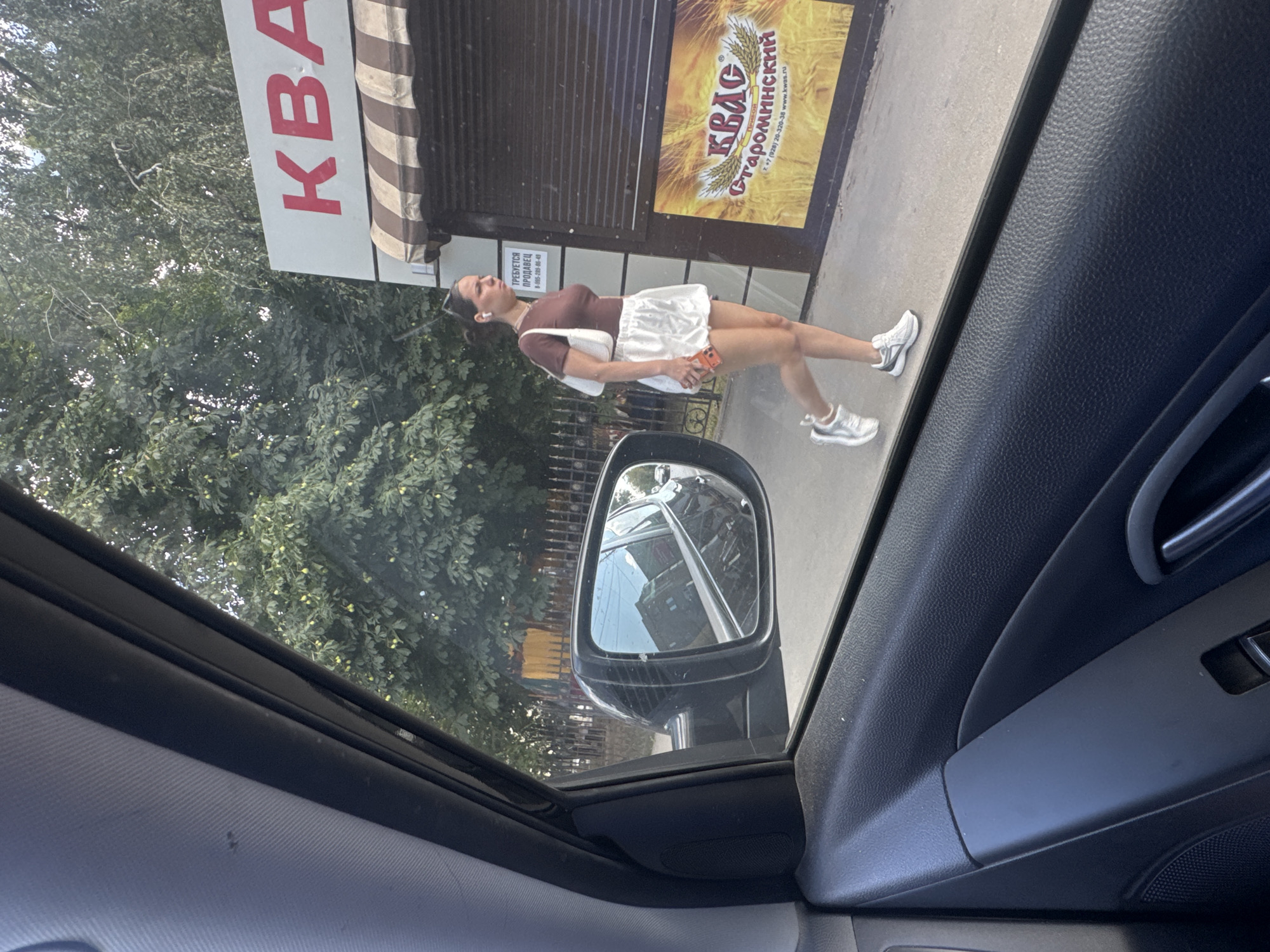 The image shows a woman walking on a sidewalk in front of a building with a sign that reads "КВАС" (Kvass). The building appears to be a kiosk or small shop. The woman is wearing a brown top, white shorts, and white sneakers, and carrying a white purse and a cell phone. The scene is viewed from inside a car, with a side mirror reflecting buildings and a street. Trees and a fence are visible in the background. The signage on the building and the reflection in the mirror suggest the location is likely Rostov-on-Don, Russia.