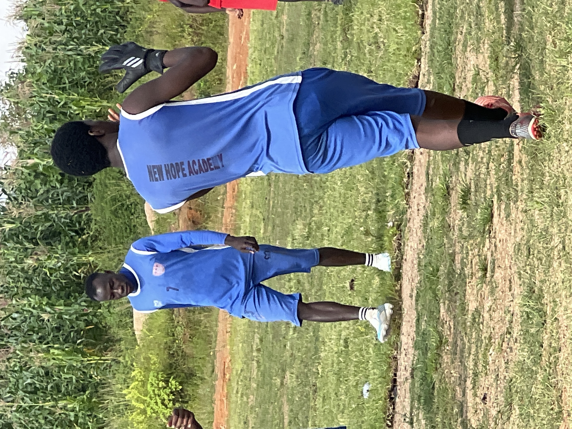 The image depicts a soccer field in Jos, Nigeria, with two young men in blue uniforms. The man on the left is wearing a long-sleeved blue jersey with the number "7" and a crest on the chest. He also has blue shorts and white socks with black stripes. The man on the right is wearing a sleeveless blue jersey with "NEW HOPE ACADEMY" written vertically on the back. He has blue shorts, black socks, and black goalkeeper gloves. He is facing away from the camera, with his right arm raised. The background shows a dense field of tall green plants, likely corn stalks, and dry, brown earth with sparse grass. The overall lighting suggests it is daytime.