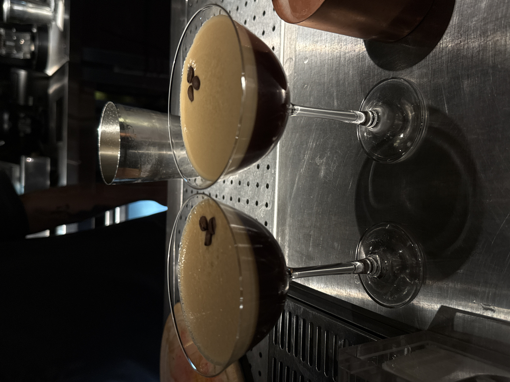 The image shows two espresso martinis in martini glasses, garnished with three coffee beans each, sitting on a stainless steel surface in a dim, indoor setting. The surface appears to be part of a bar or countertop, with equipment and a drink shaker visible in the background. The exact location in Yerevan, Armenia cannot be determined from the image. No people are visible in the immediate foreground, although a hand is partially visible on the left side of the image.