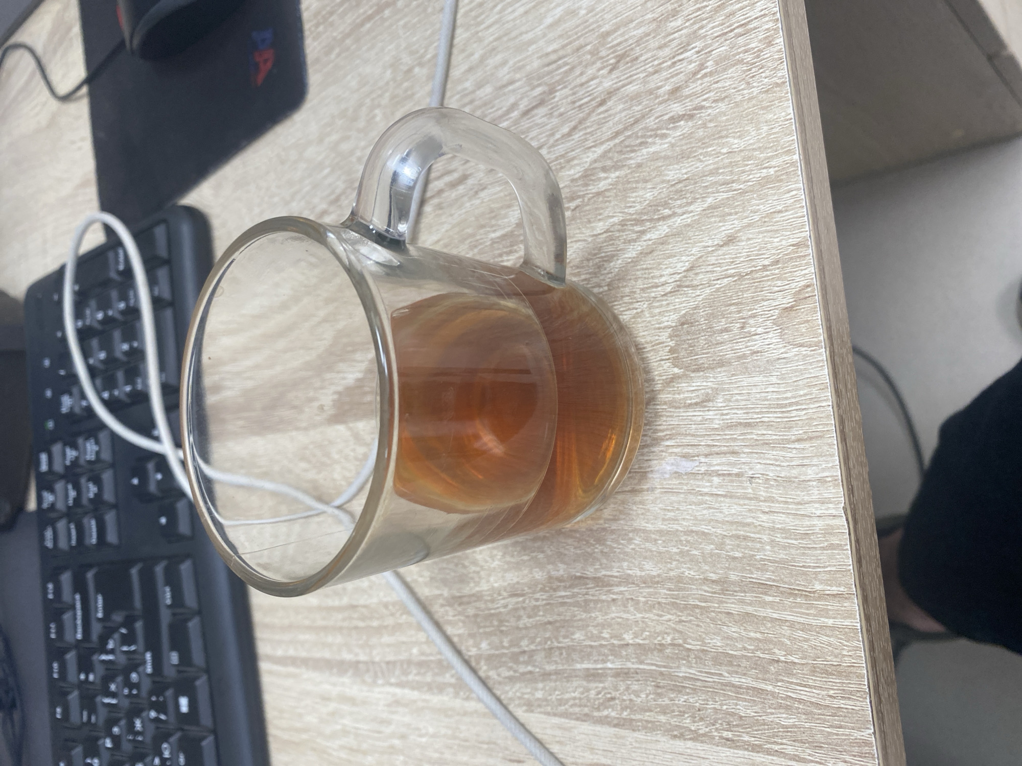 A clear glass mug filled with amber liquid, likely tea or coffee, sits on a light wood-grain surface. The mug has a transparent handle and the liquid is about halfway full, with reflections visible on the glass. In the background, a black keyboard is partially visible, with a white cable running from it. To the left of the keyboard is a black mousepad with a logo that includes red and blue elements. The overall scene appears to be a desk or workspace, with a subtle hint of a shelf or furniture edge on the right side of the image. The lighting suggests it could be daytime. No people are directly visible, but the presence of a keyboard and mouse implies a person was recently using or is about to use them. The setting appears to be indoors, likely an office or home environment. No visible text is legible in the image.