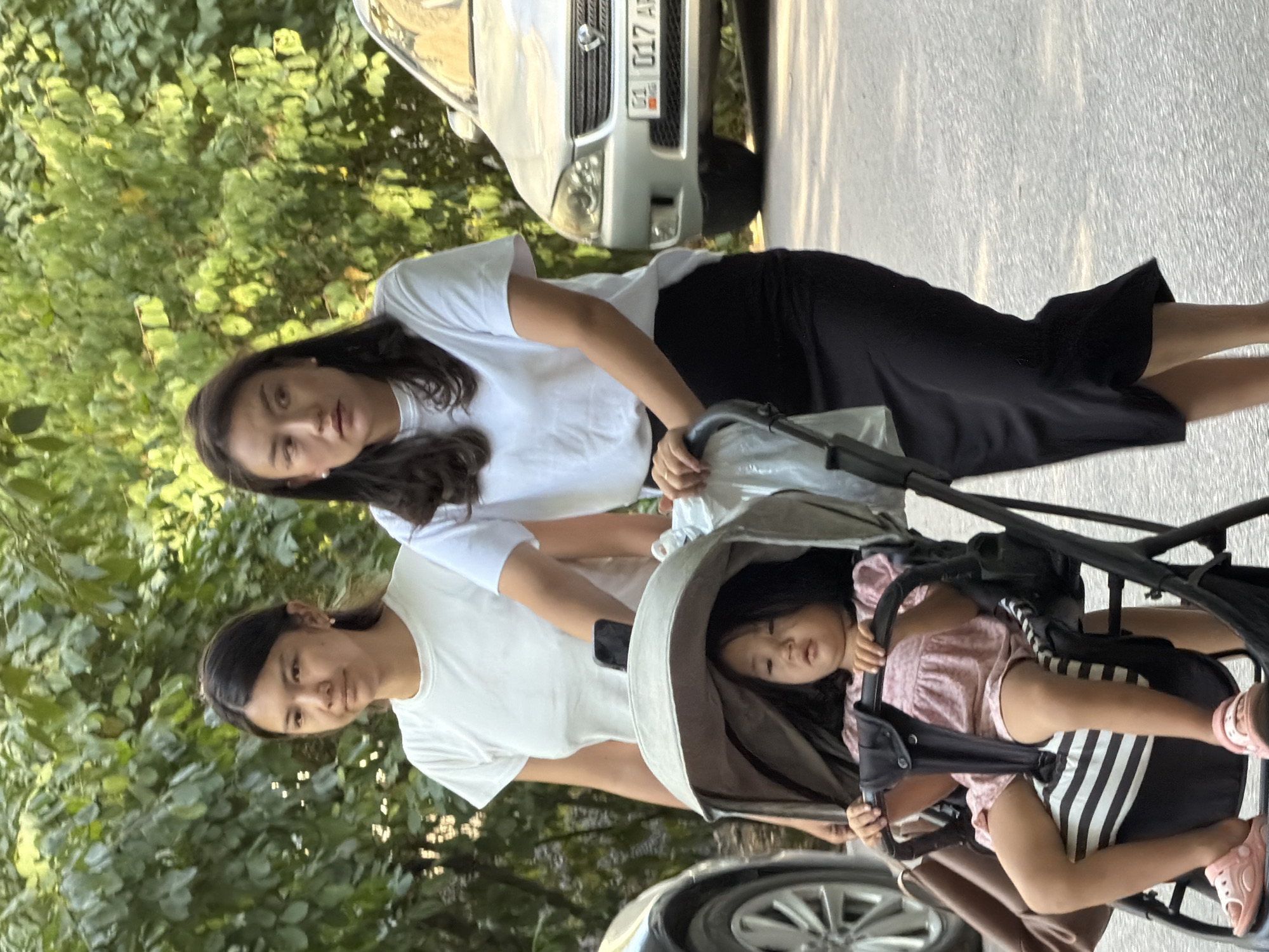 Two women and a young child are outdoors on a paved surface, possibly a sidewalk or street, with lush green foliage in the background. The woman on the right, wearing a white t-shirt and a black skirt, is pushing a stroller with a young child in it. The child, wearing a pink dress and pink sandals, is seated in the stroller, looking forward with a neutral expression. The woman on the left, wearing a white t-shirt, stands behind the stroller, looking towards the left. Behind the women and stroller, a silver sedan is parked. The lighting suggests it is daytime, possibly late afternoon, with some sunlight filtering through the trees.