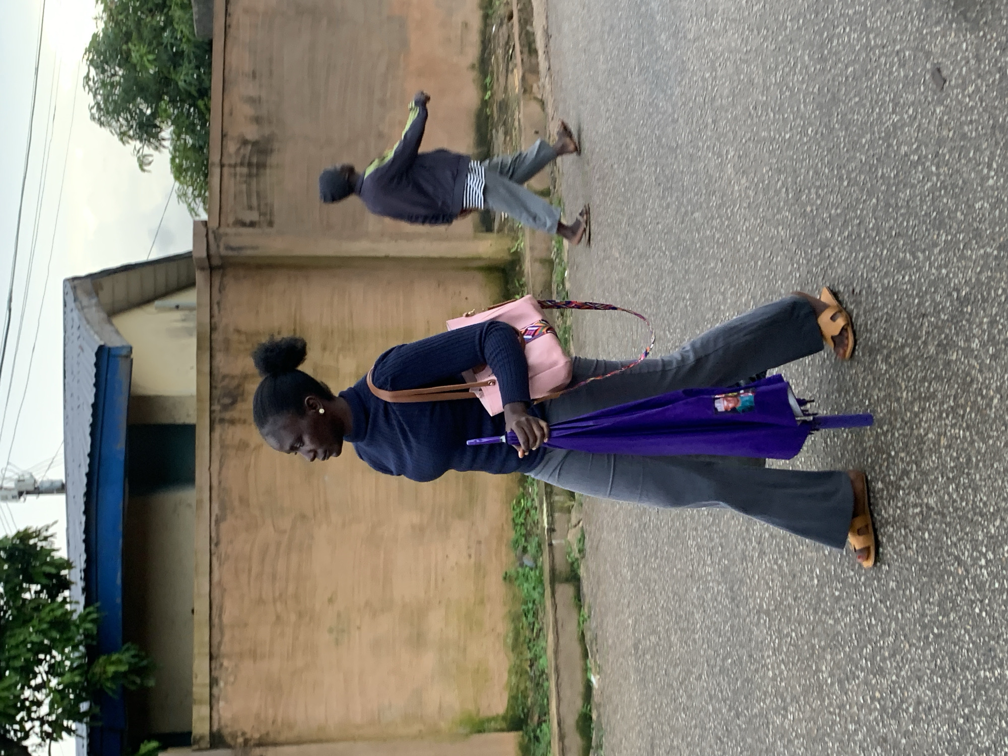 In Jos, Nigeria, on an overcast day, a young Black woman walks past a beige wall. She has her hair pulled back in a bun and is wearing a dark blue turtleneck sweater, dark gray flared pants, and tan sandals. She carries a pink purse over her right shoulder and a closed purple umbrella in her left hand. The umbrella has a picture of a man on it. In the background, another person walks away from the camera, wearing gray pants and a dark jacket with green stripes on the sleeves. The scene is on a paved street in front of a building with a dark roof. Green foliage is visible along the wall and on the left side of the image.