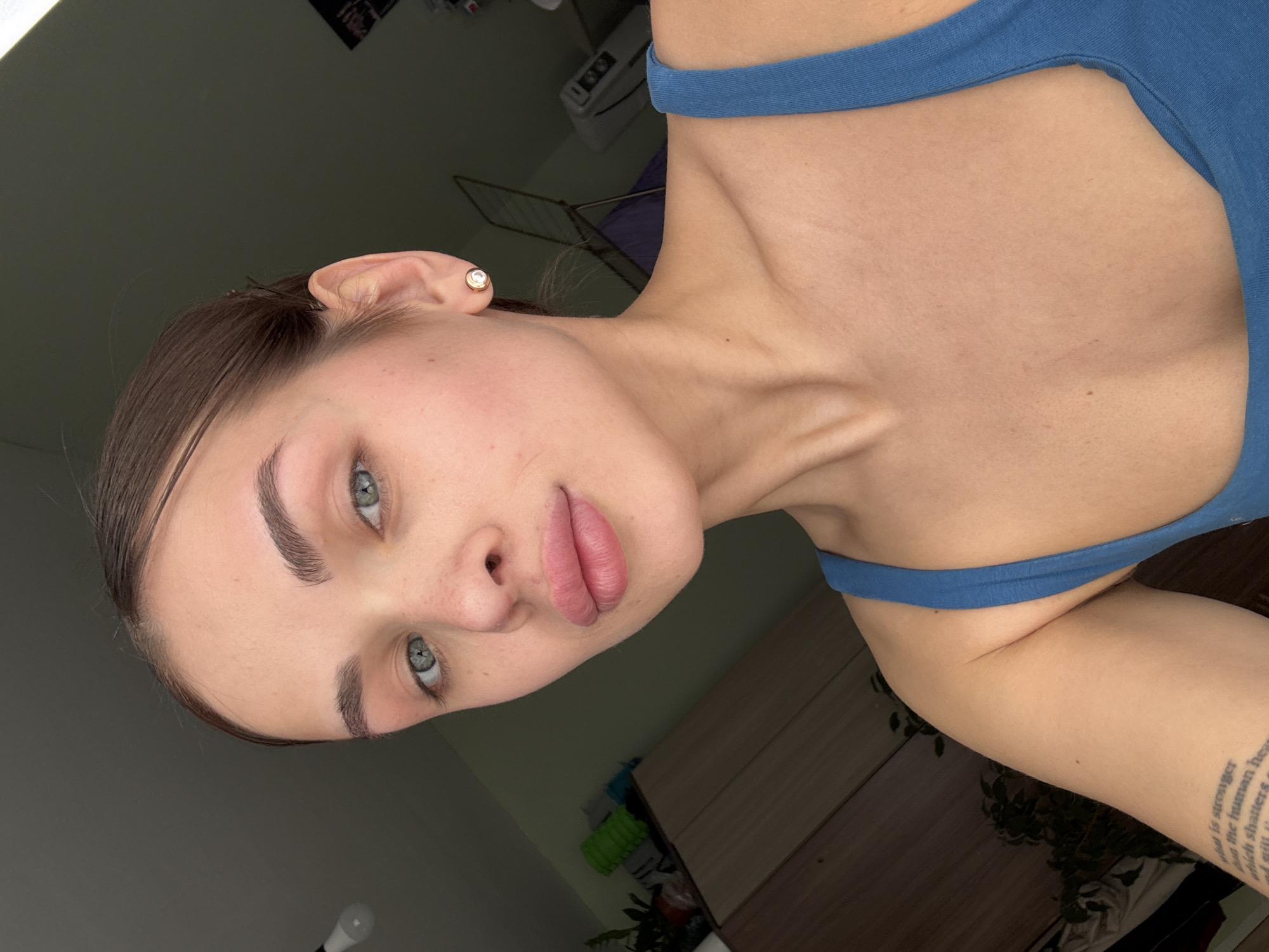 A young woman with light skin, brown hair pulled back, and light green eyes is photographed indoors. She wears a blue tank top. Her left ear is visible with a stud earring. Tattoos are visible on her left arm. The background includes a wall, furniture, and a plant. The location is not identifiable as Sarapul, Russia from the image.