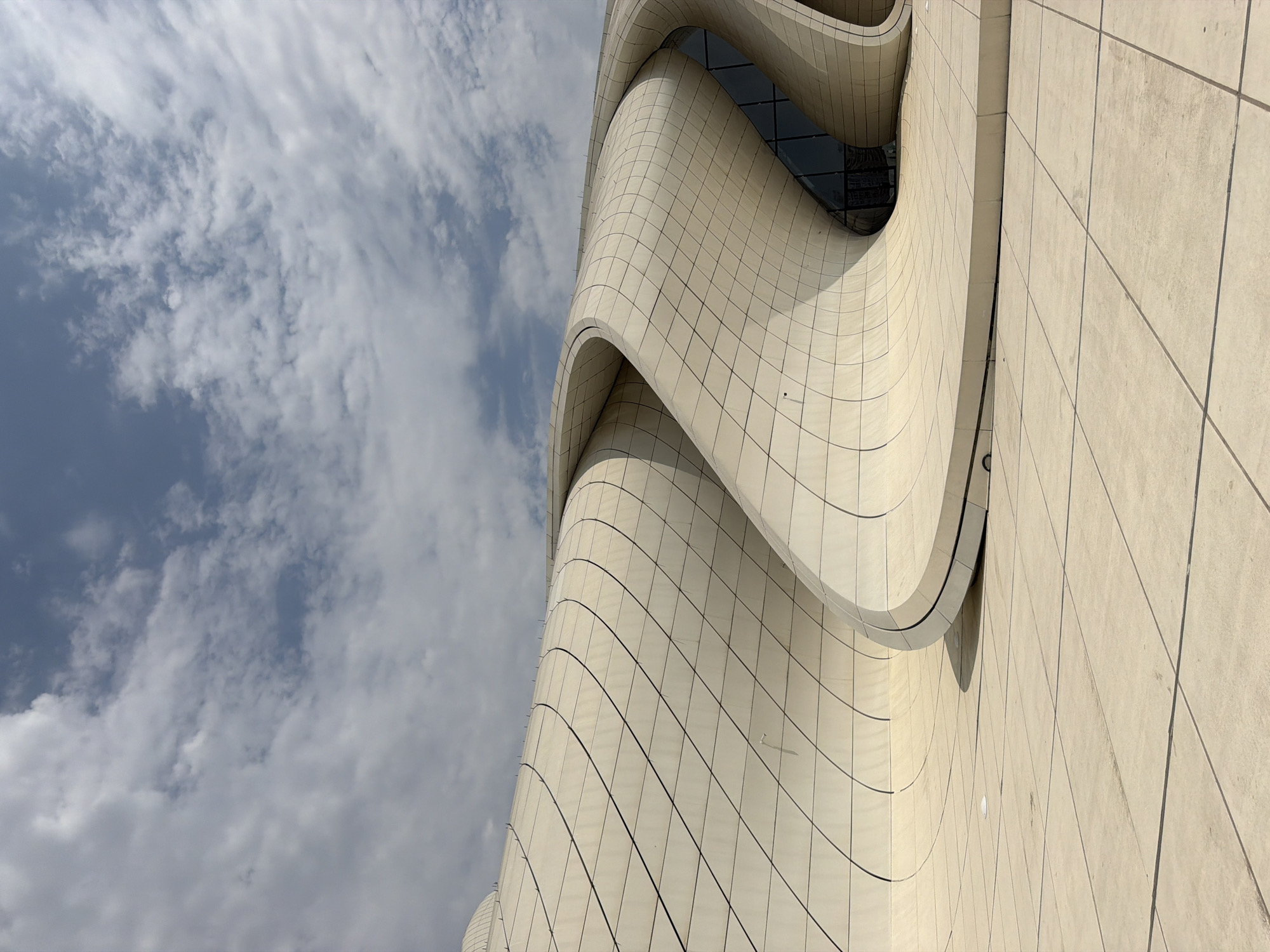 The image shows the Heydar Aliyev Center in Baku, Azerbaijan, on a partly cloudy day. The building's exterior features flowing, curved white panels with a grid pattern. The foreground is a paved plaza with a similar grid pattern. The sky above is filled with white and gray clouds against a blue sky. There are no people or animals visible in the frame. The architecture is modern and avant-garde, with smooth, undulating surfaces and large glass elements integrated into the curves.