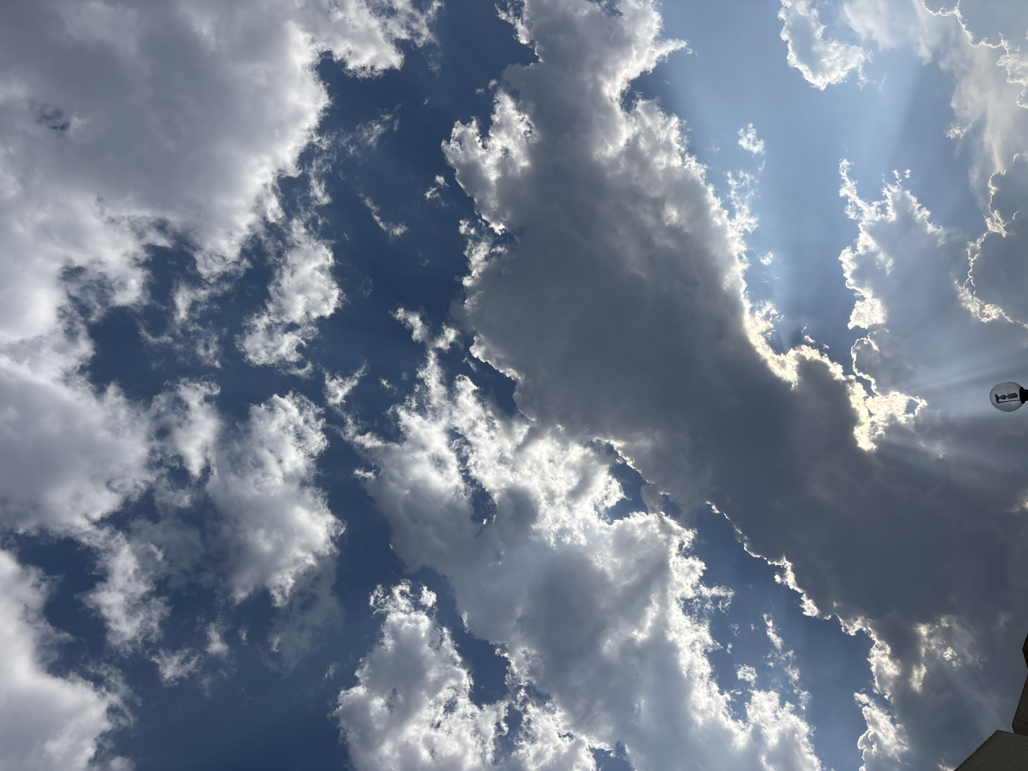 The image depicts a sky filled with a mixture of cumulus and cirrus clouds, with varying shades of white and gray. Patches of blue sky are visible between the clouds. Sunlight streams through gaps in the cloud cover, creating visible rays. A portion of a lamppost is visible in the top right corner.
While I can describe the image, I cannot confirm the location as Puebla, Mexico based solely on the visual information. Additional contextual information would be required for that.