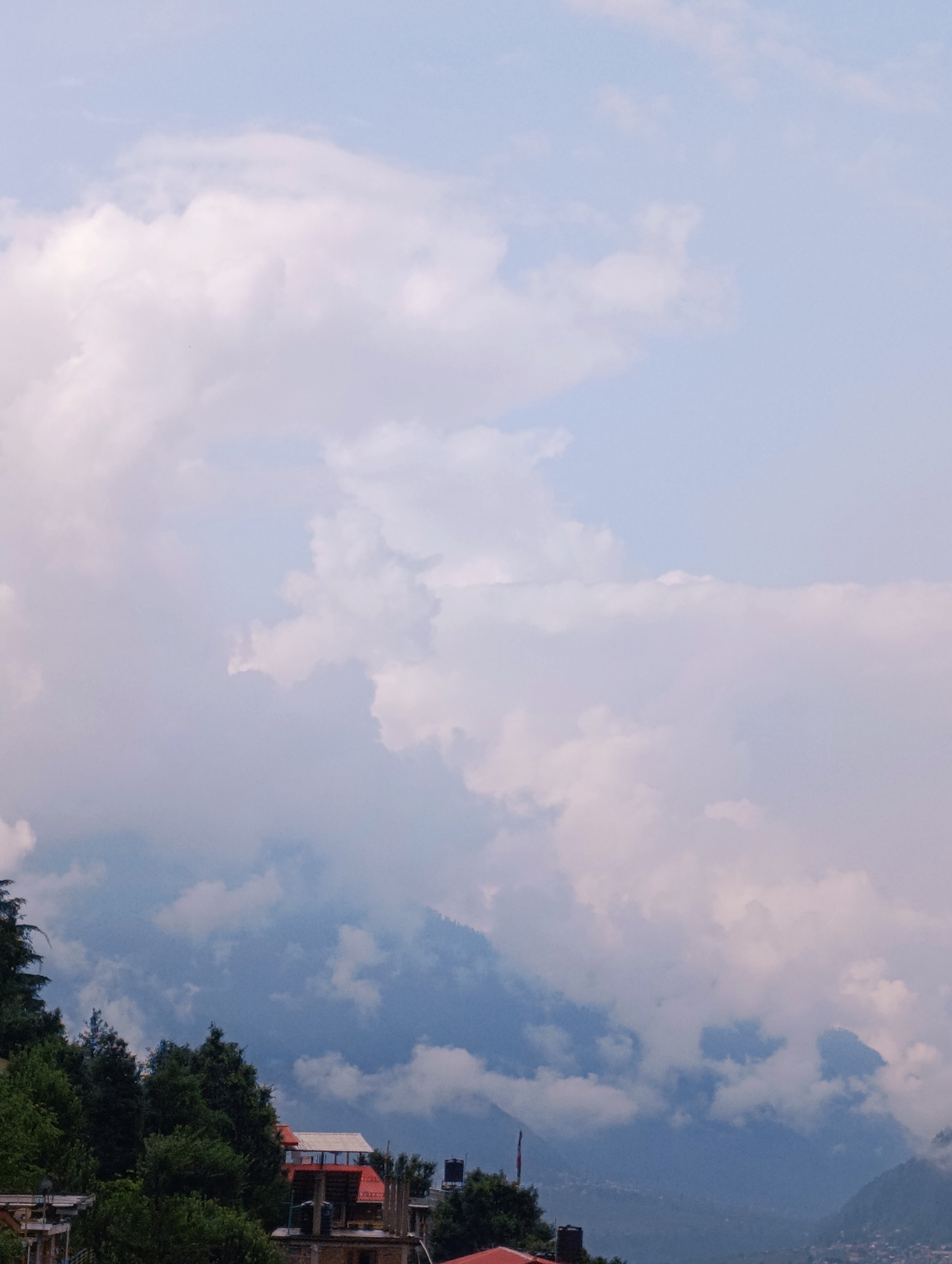 A high-angle, outdoor shot shows a misty mountain landscape with clouds partially obscuring the peaks and trees. In the foreground, a few buildings with red roofs and water tanks are visible amongst dense green trees, suggesting a settlement nestled within the hilly terrain. The sky is a pale blue, filled with soft, white and grey clouds that create a diffused lighting effect over the scene. The overall atmosphere is serene and slightly overcast, typical of a mountain environment.