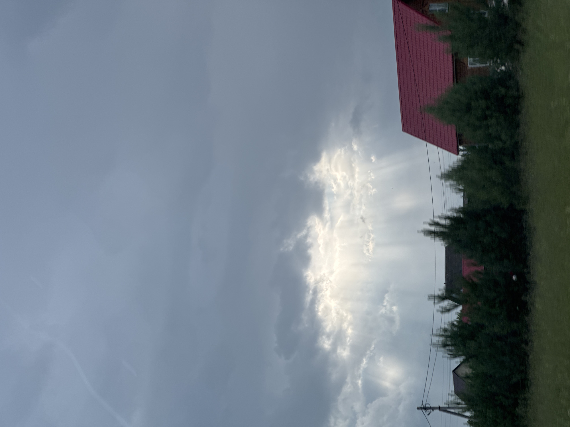The image shows a landscape with a cloudy sky and houses with red roofs, surrounded by trees. The sun's rays are breaking through the clouds, creating a dramatic effect. The scene appears to be set in a rural area, possibly Kuyeda, Russia, given the provided location context. The time of day is likely late afternoon or evening, as indicated by the low angle of the sun and the diffused light. The weather appears to be unsettled, with a mix of heavy clouds and bright sunshine. The overall mood is one of natural beauty and perhaps a touch of drama, with the interplay of light and shadow. There is a power line visible in the foreground, running horizontally across the image. The houses are typical of rural Russian architecture, with gabled roofs and wooden siding. The trees are dense and green, suggesting it is a warm season. No people are visible in the image. There is no visible text.